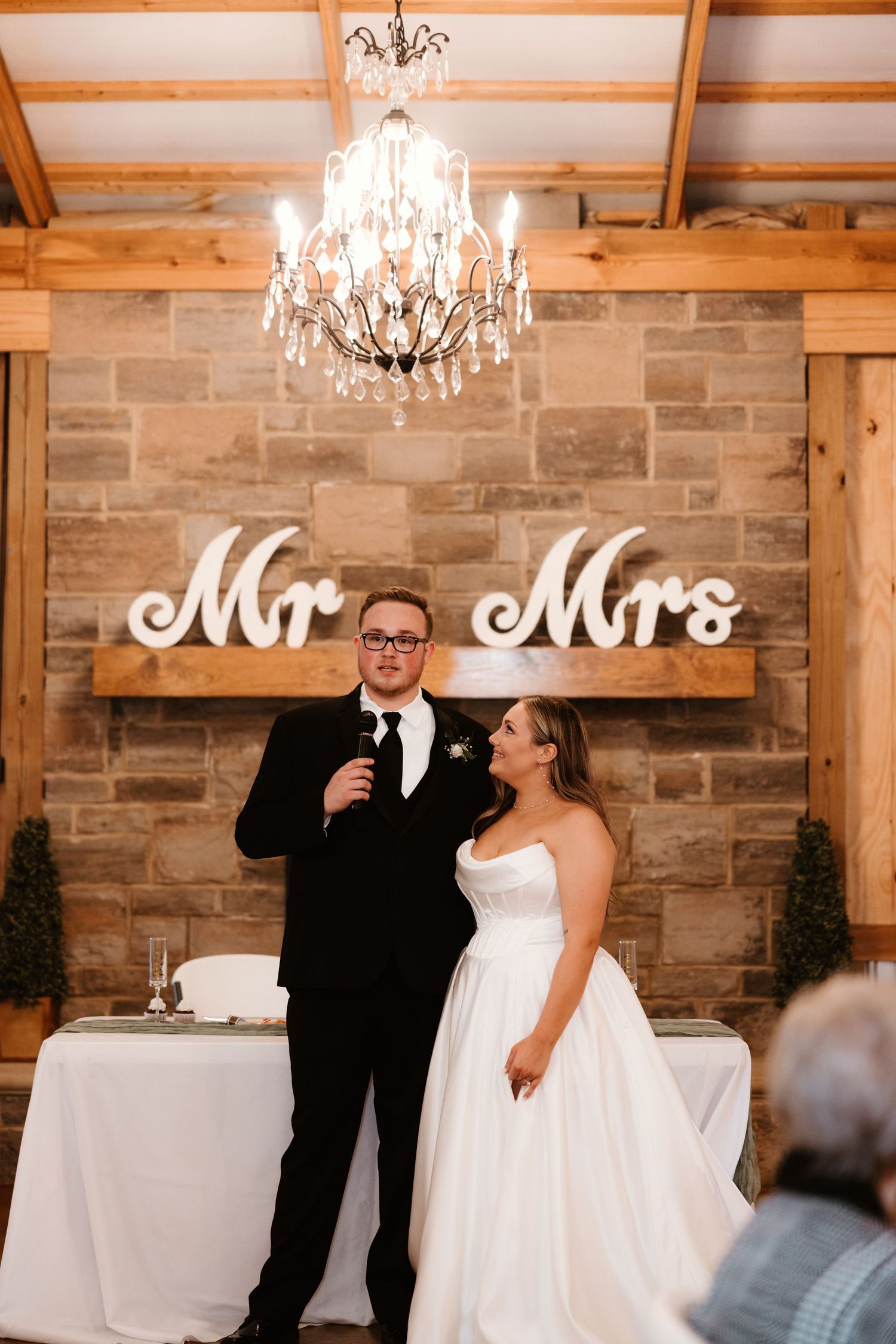 Elegant wedding reception venue with crystal chandelier and 'Mr & Mrs' sign on rustic stone wall backdrop.