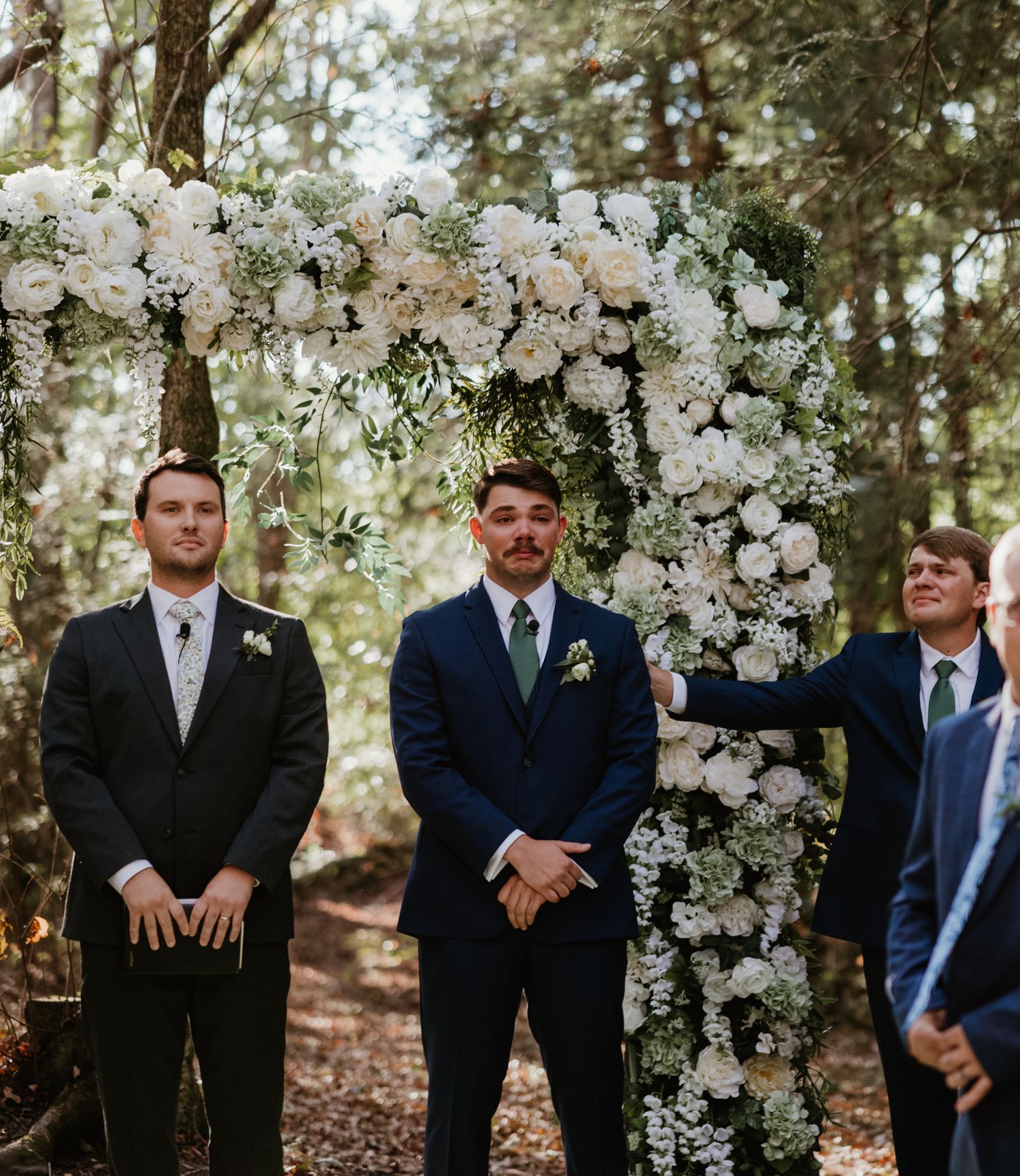 Groomsmen standing at a white floral wedding arch in a wooded setting.