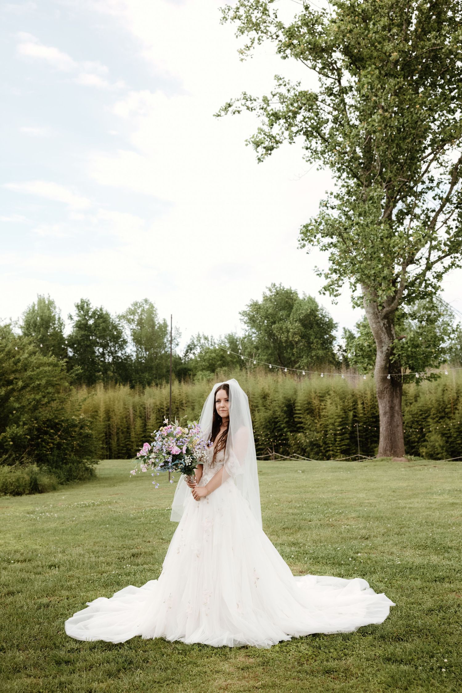 A bride in a flowing white wedding gown with train holds a colorful bouquet in an outdoor garden setting.