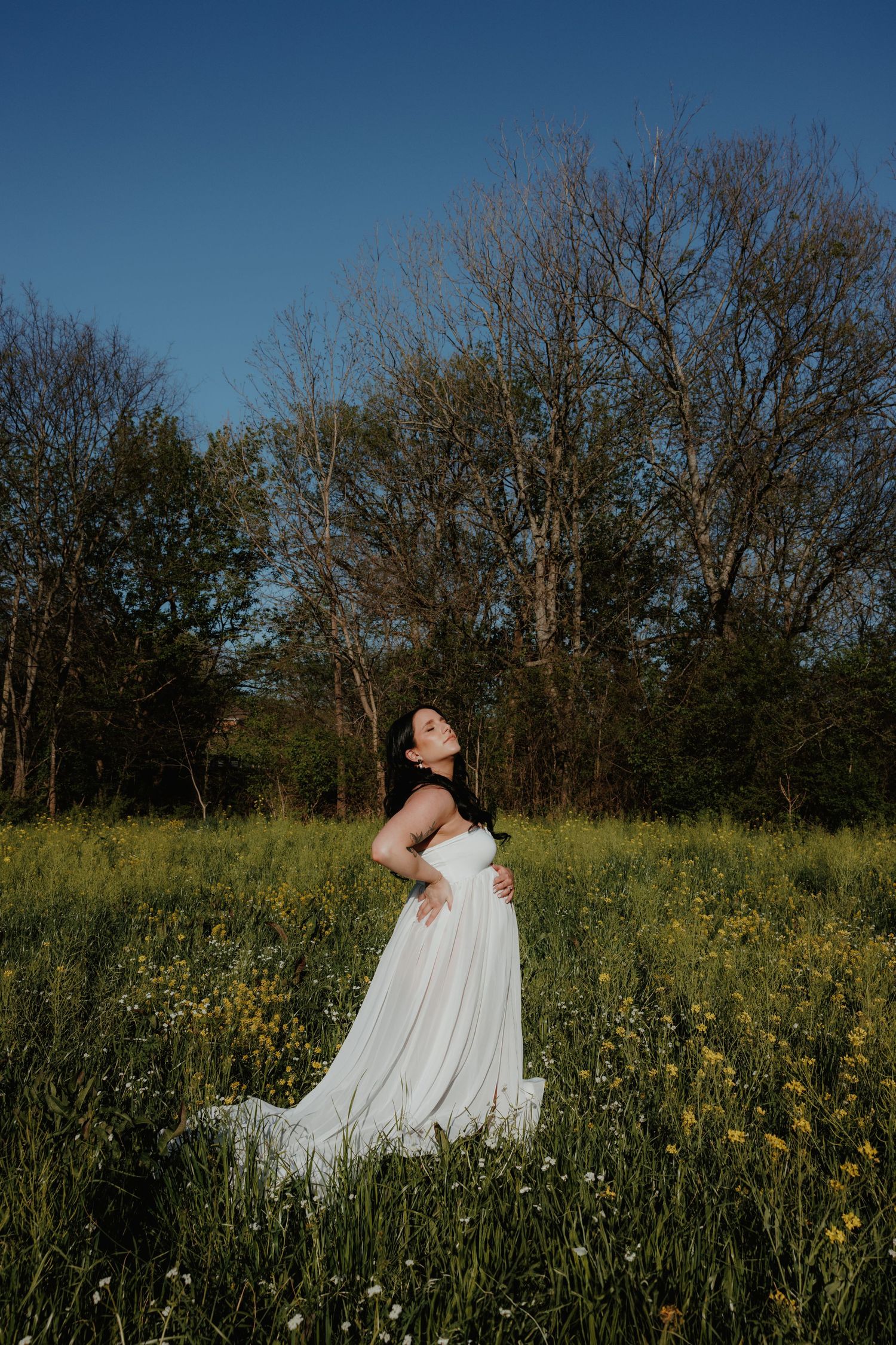 A maternity photo in a wildflower field with trees and blue sky in the background.