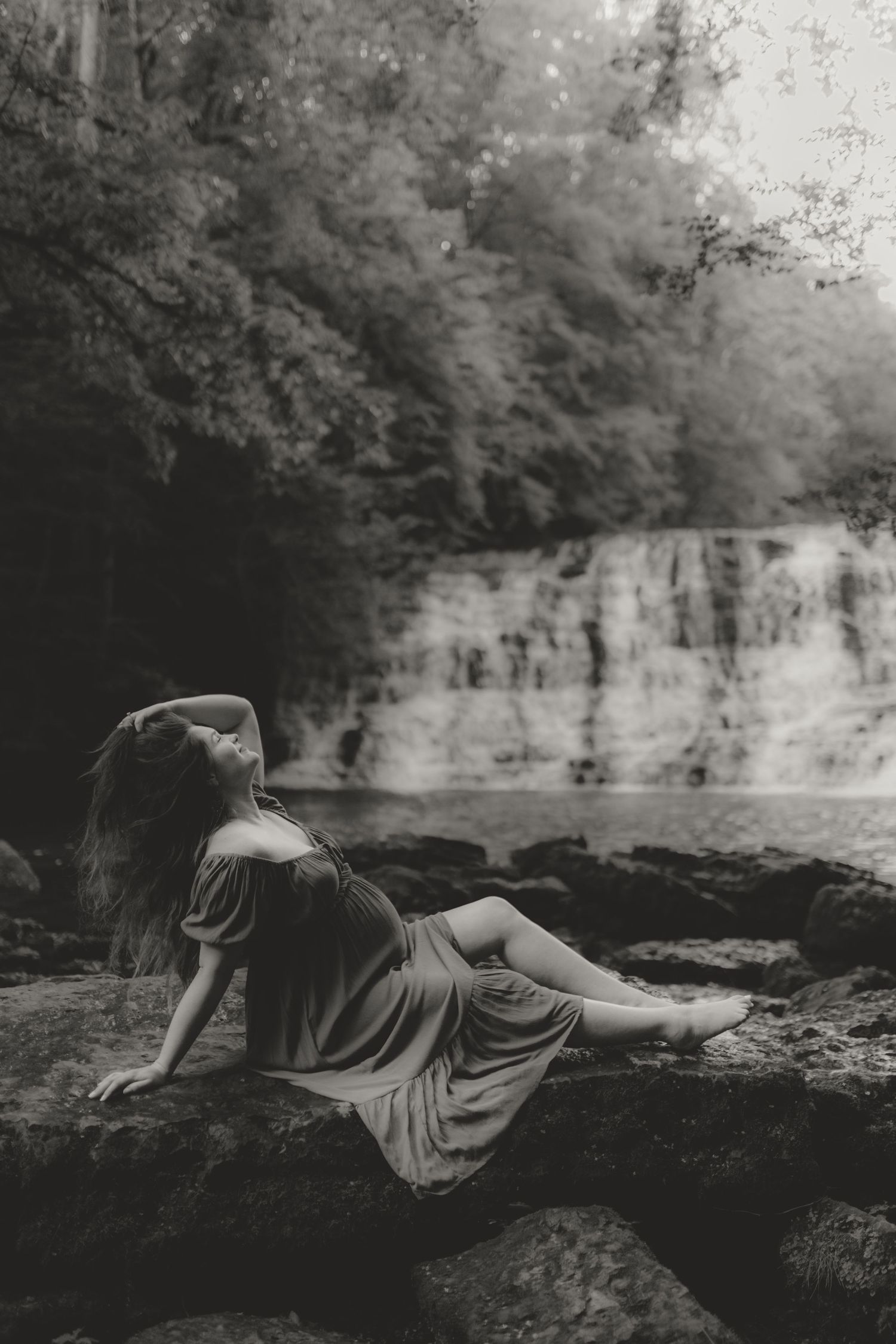 Artistic black and white portrait of a figure reclining on rocks near a waterfall.