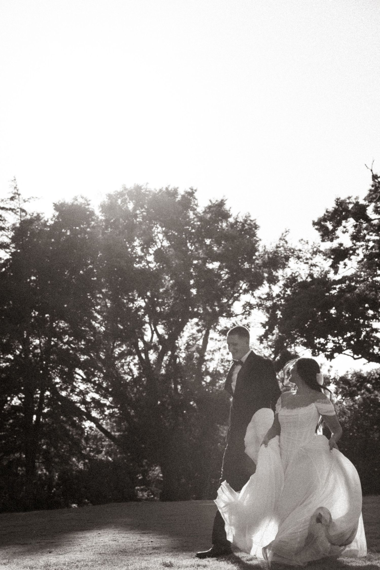 Black and white photo of a bride and groom walking outdoors among trees, her gown flowing gracefully in the breeze.