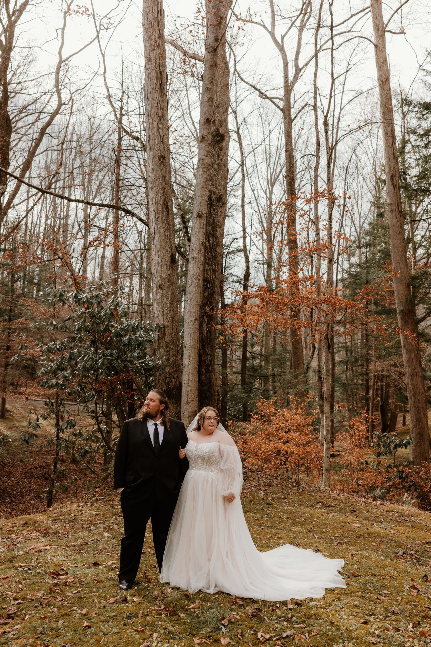 Bride and groom stand in an autumn forest, she in a flowing white gown with veil, he in a black tuxedo.