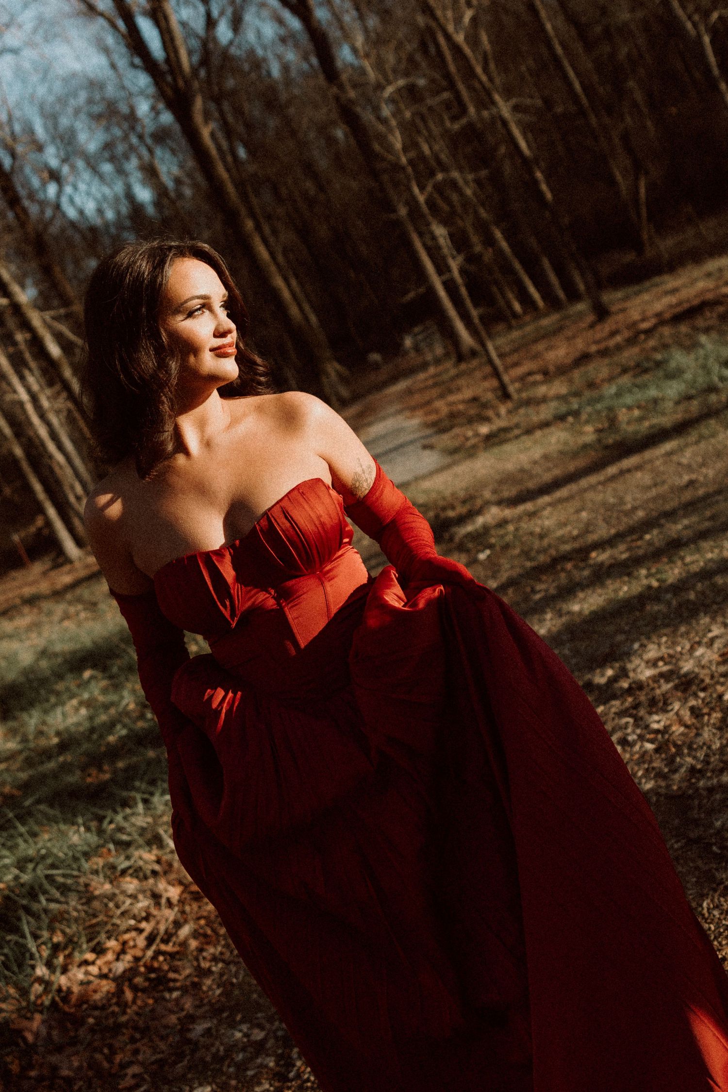 Woman in elegant red strapless gown with gloves poses gracefully in a sunlit autumn forest setting.