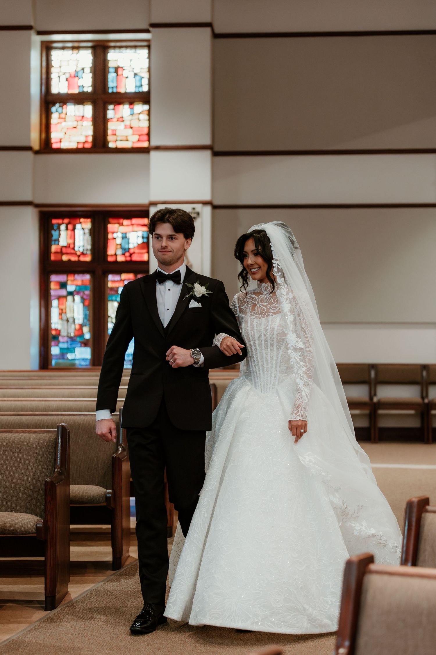 Bride and groom walking down church aisle in elegant wedding attire with stained glass windows backdrop.