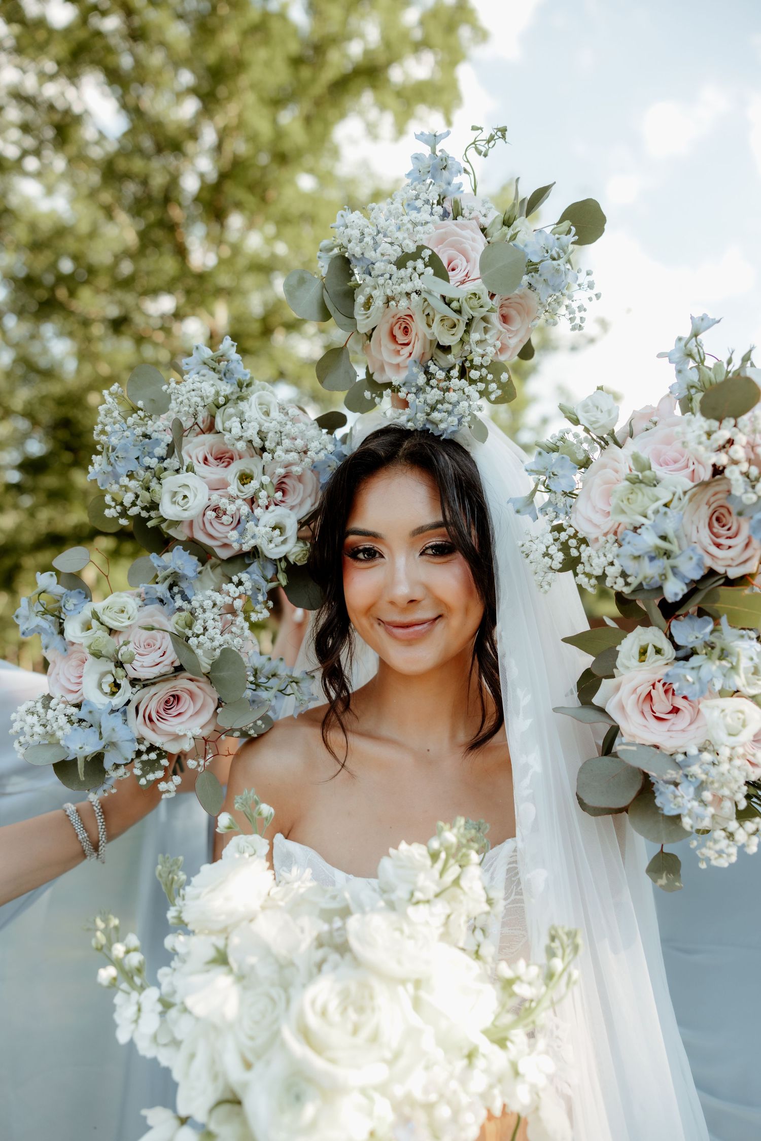 Beautiful bride surrounded by bridesmaids holding pink and white rose bouquets outdoors on wedding day.