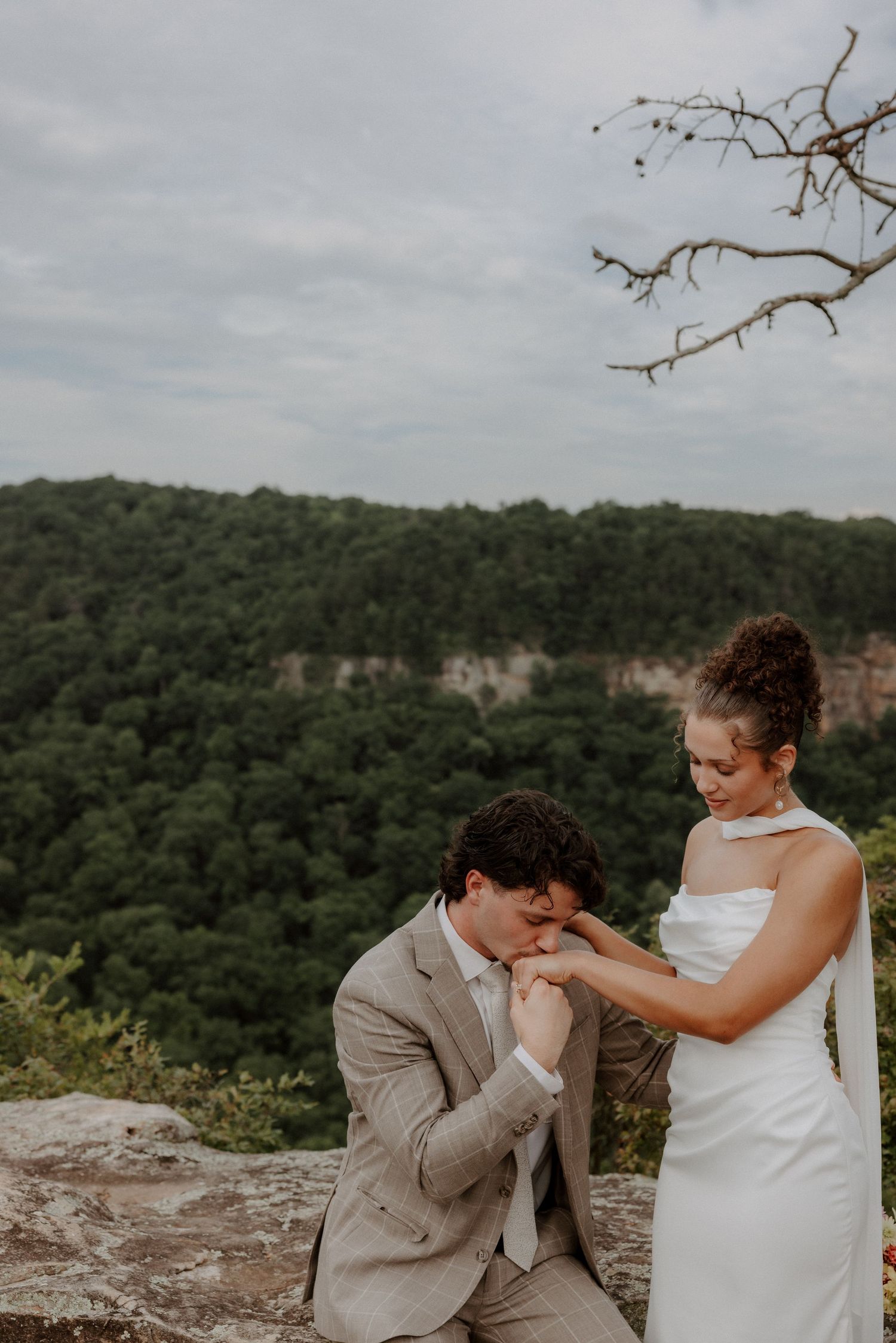 Groom kneeling to kiss bride's hand on rocky overlook with lush forest backdrop.