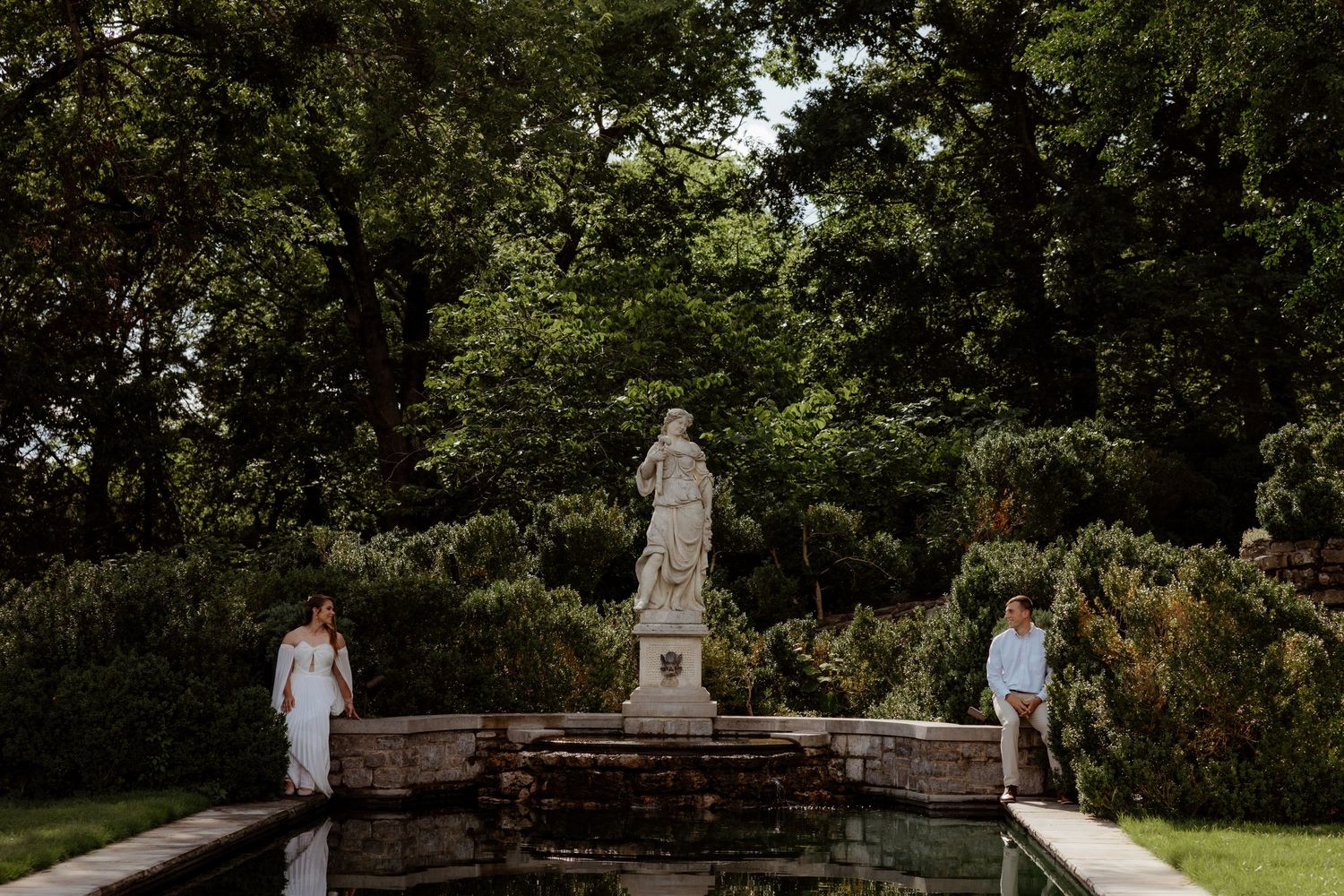 Couple standing beside a stone fountain with classical statue in a lush garden setting.