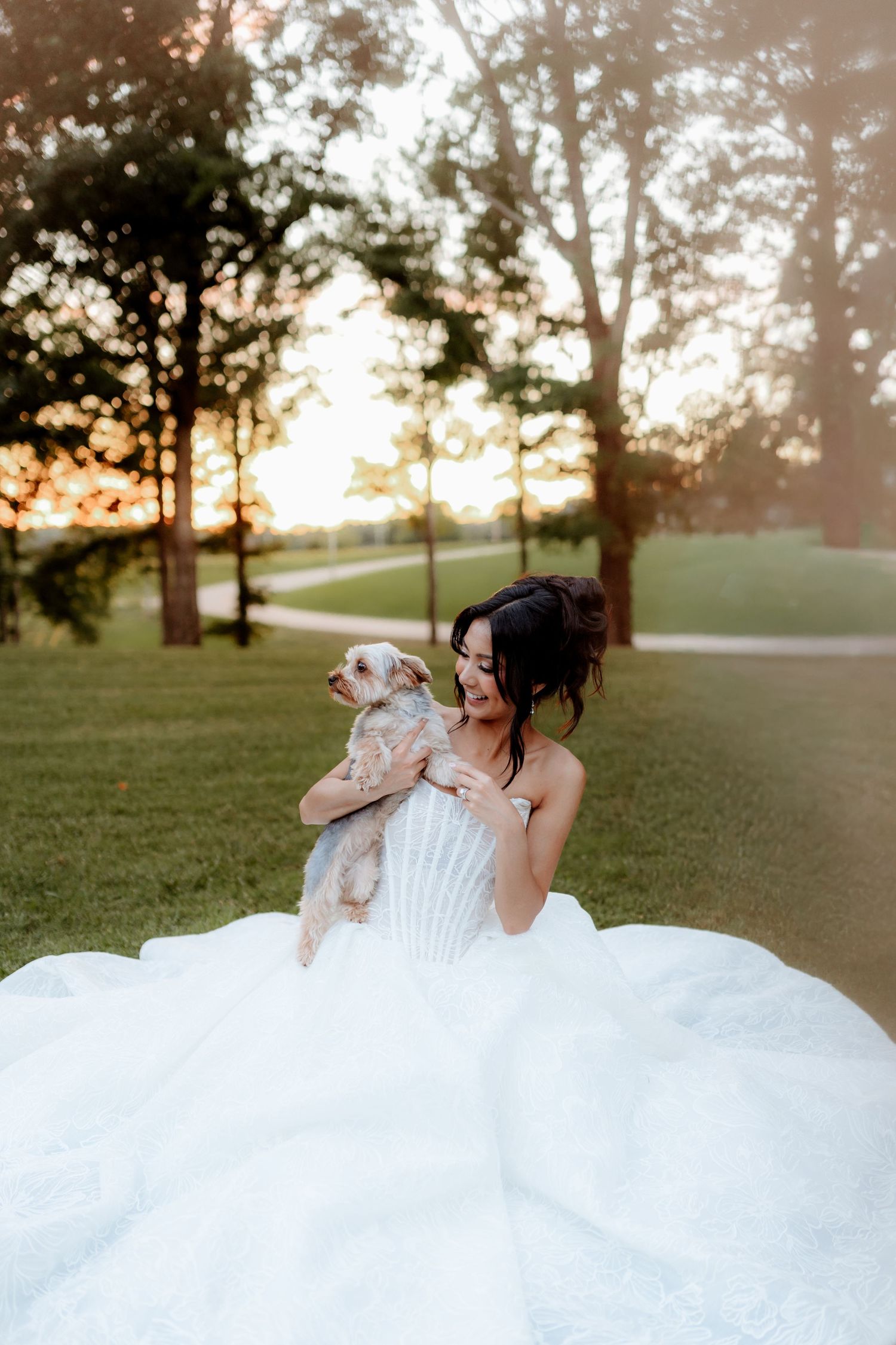 Bride in a white ballgown holds a small dog outdoors at sunset with trees and green grass in the background.