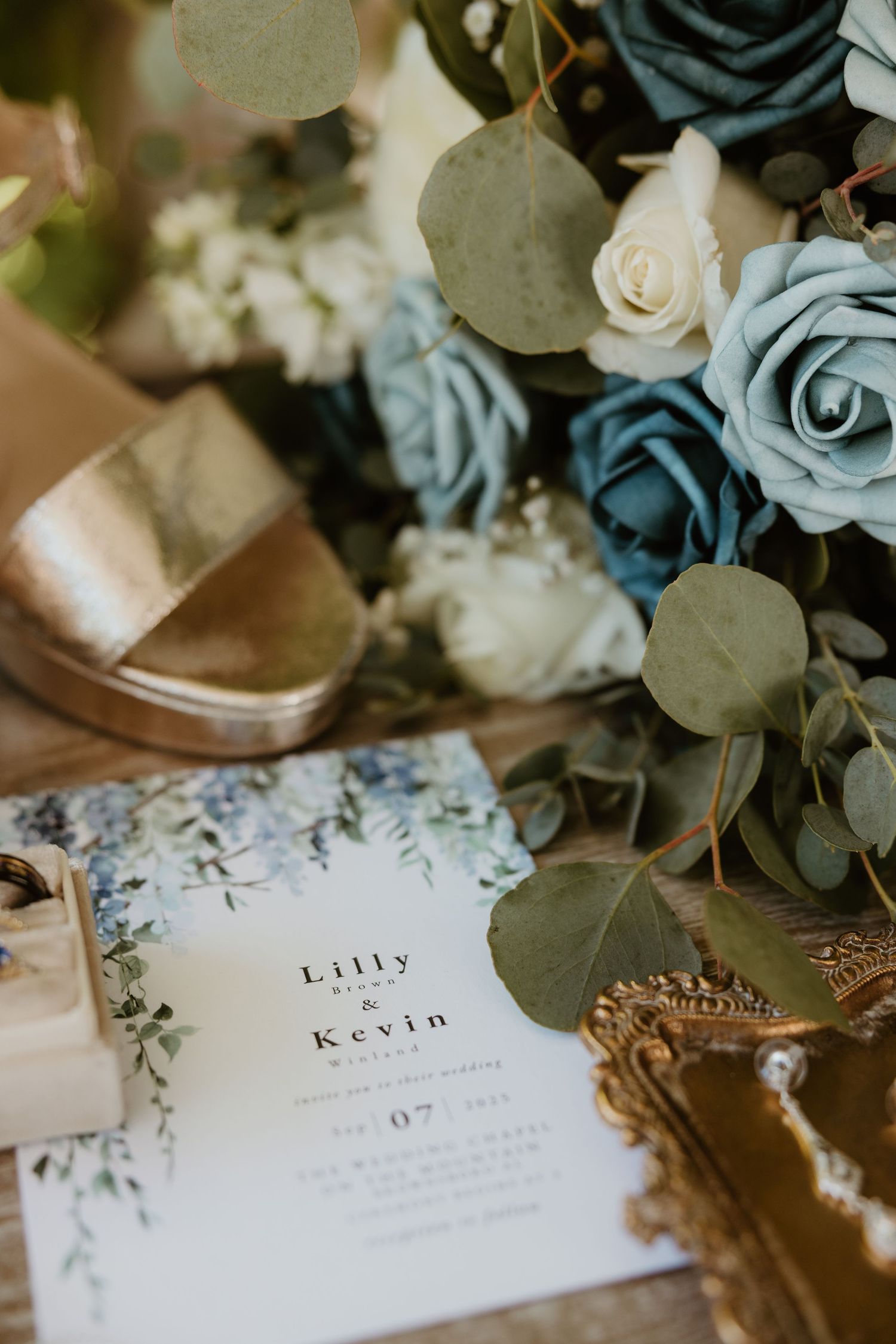 Elegant wedding stationery cards with names displayed alongside dusty blue roses and eucalyptus leaves on a white surface.