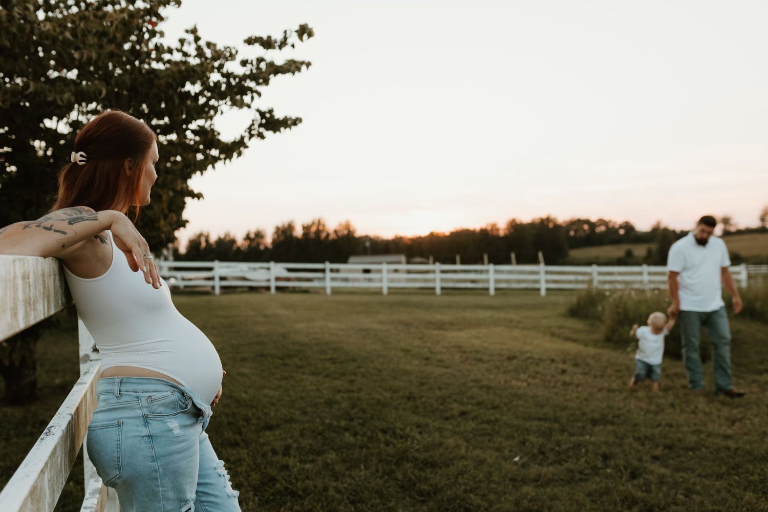 A woman leans on a white fence watching a parent and child in the distance during a peaceful countryside evening.