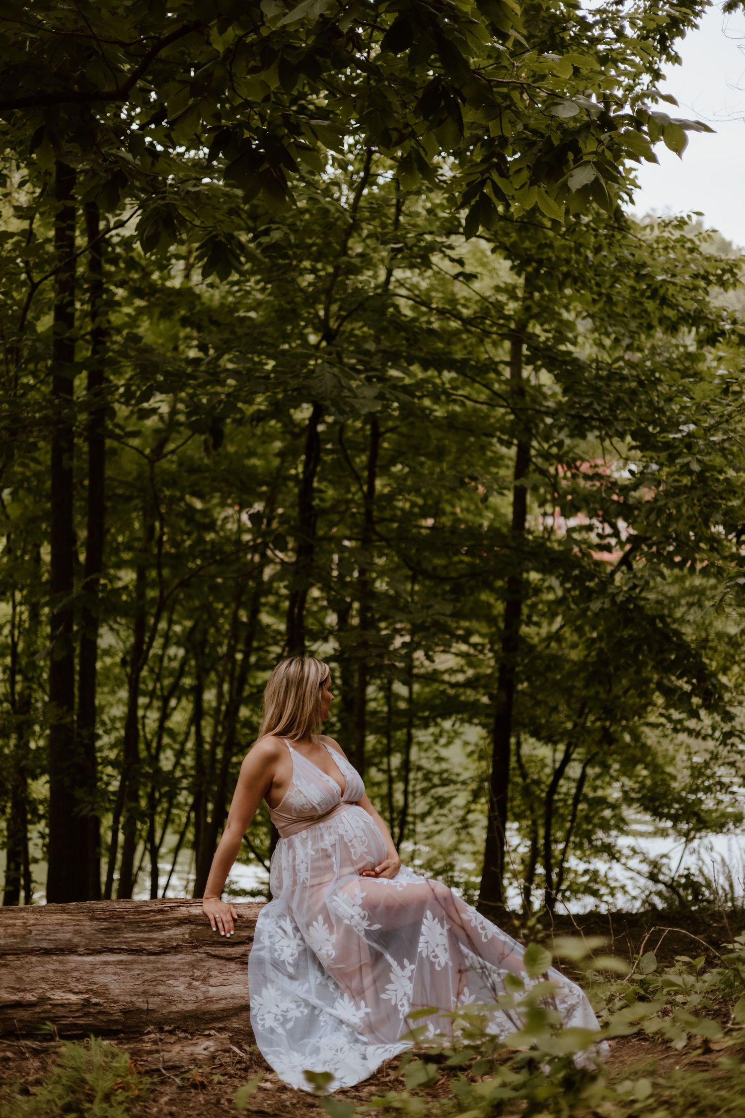 A figure in a white lace dress sits peacefully on a fallen log surrounded by lush green forest trees.