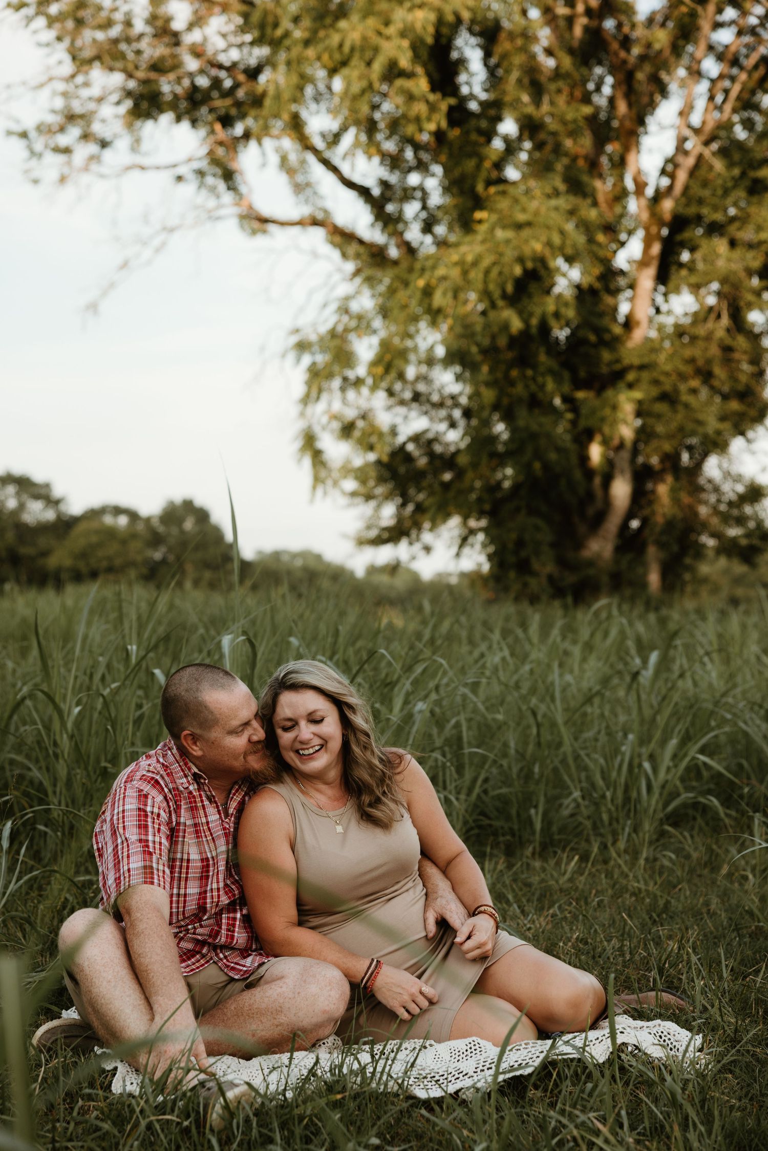 A romantic couple relaxes on a blanket in a grassy meadow under tall trees during a warm summer afternoon.