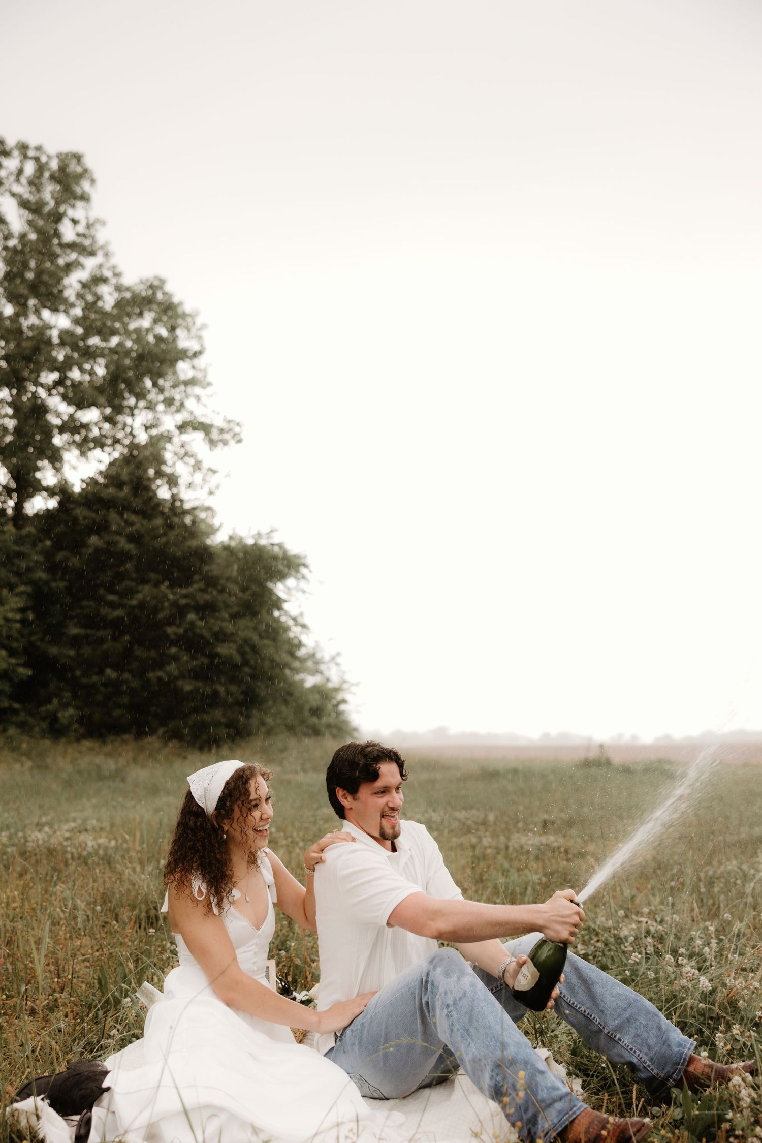 Couple spraying champagne while sitting in a grassy meadow at sunset during celebratory photoshoot.