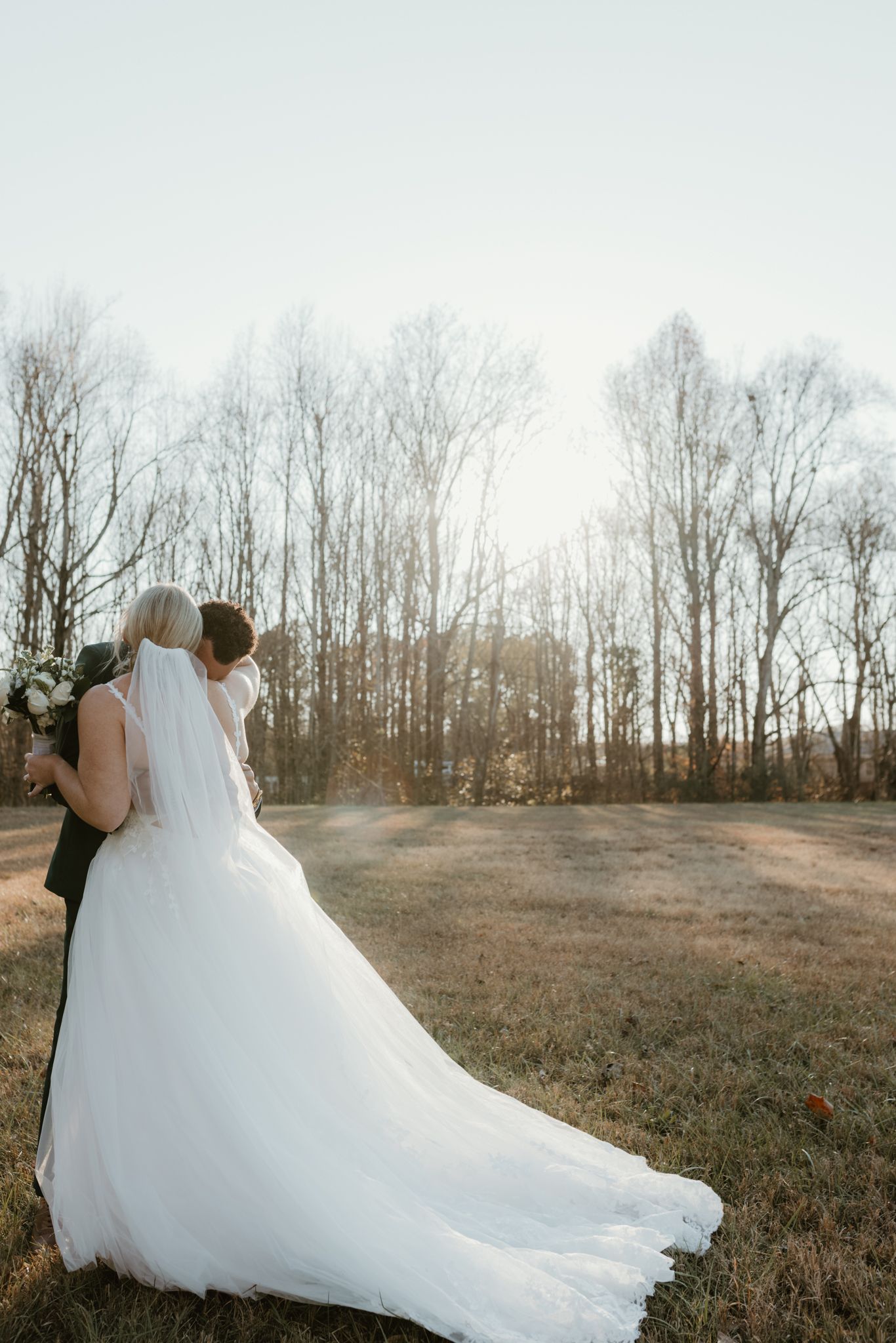 A couple in wedding attire embraces in a sunlit field with bare trees in the background.