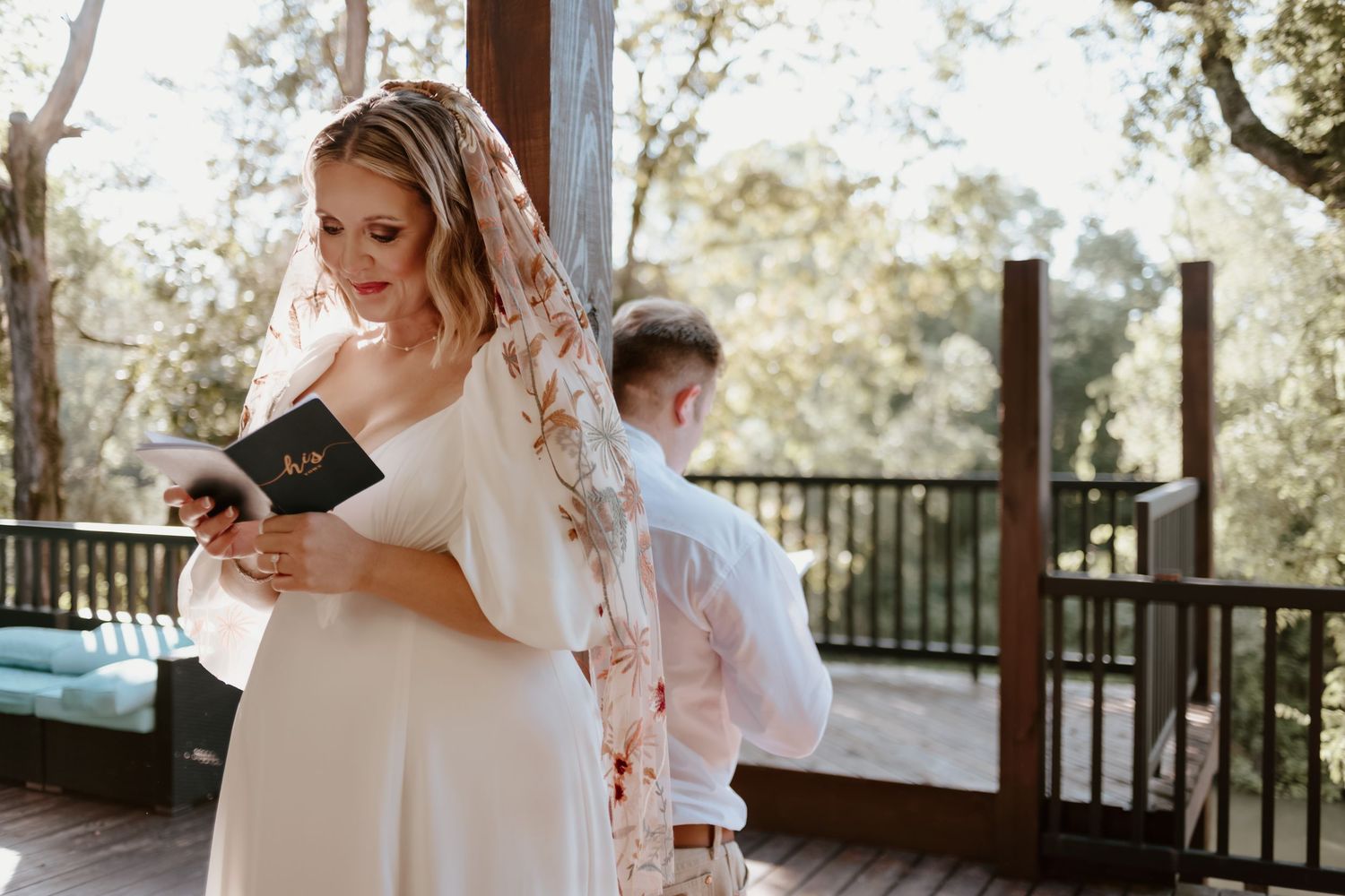 A wedding couple shares a tender embrace on a sunlit porch while photographers capture their special moment.