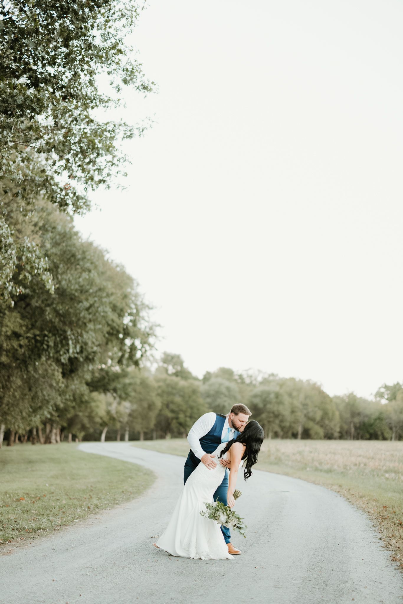 A newlywed couple shares a romantic dip pose on a country road surrounded by greenery.