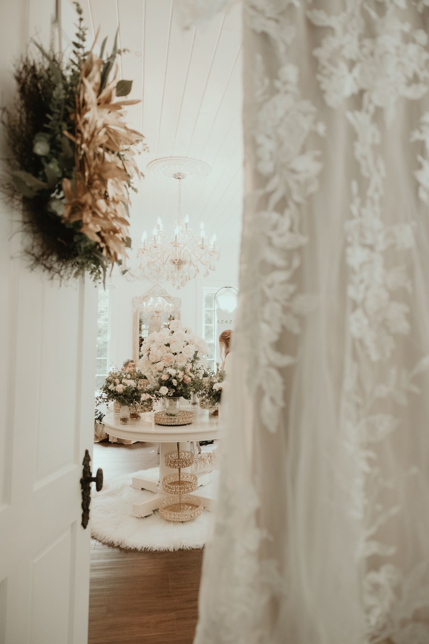 Elegant wedding cake and floral arrangement viewed through doorway with lace curtain and dried flower wall decor.