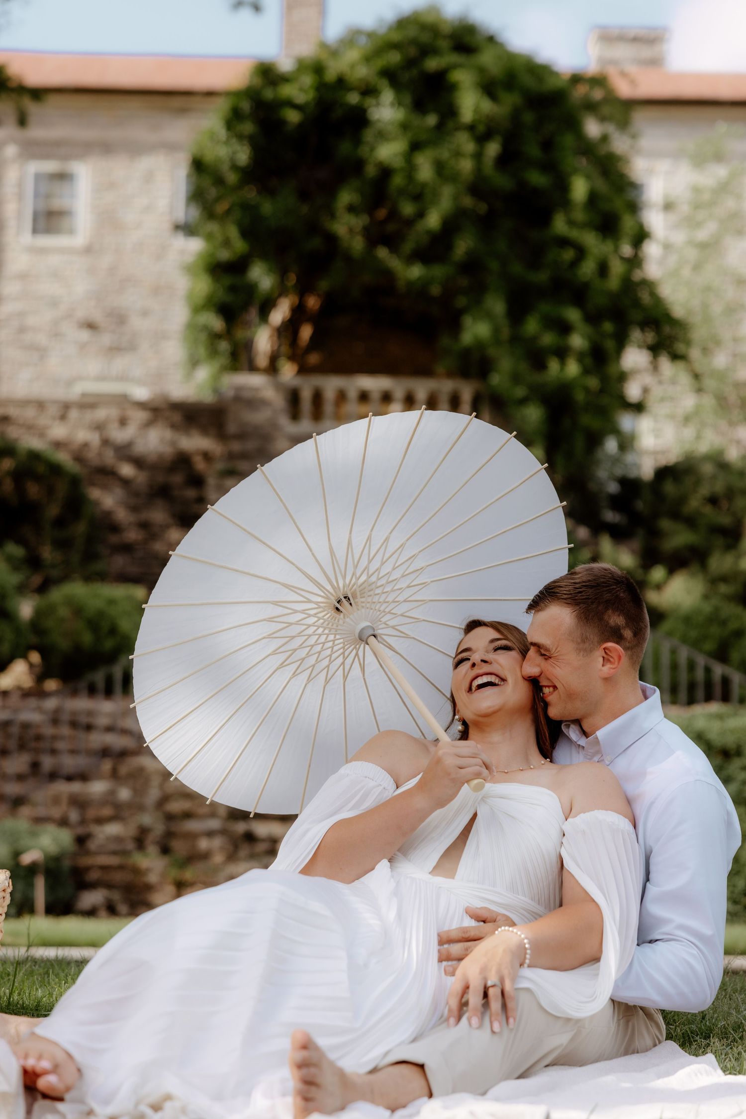 Happy couple sitting outdoors on wedding day, bride holding white parasol umbrella, groom embracing her near stone building.