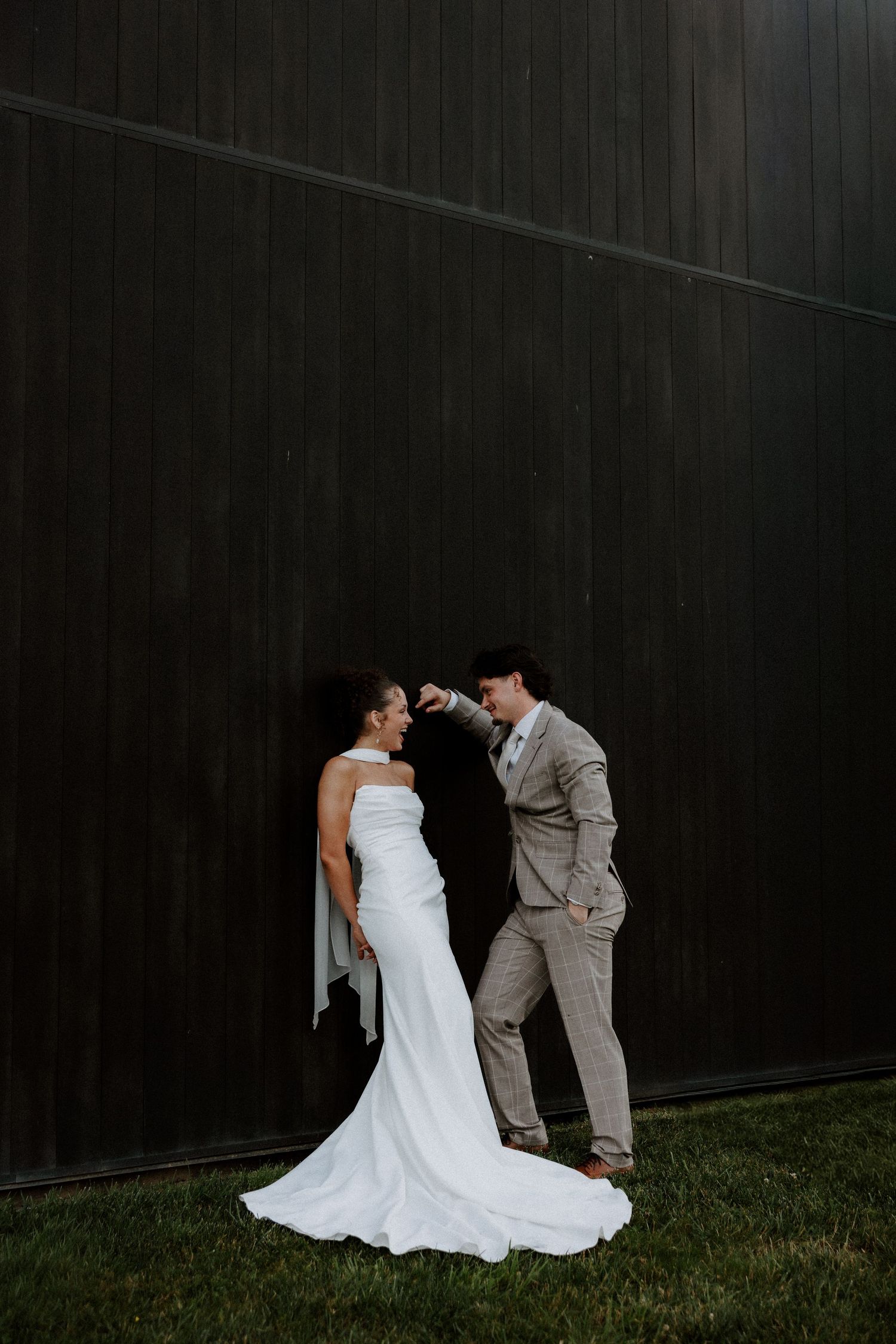 Bride and groom share a romantic moment against a dark wood wall, groom kissing her hand in elegant wedding attire.