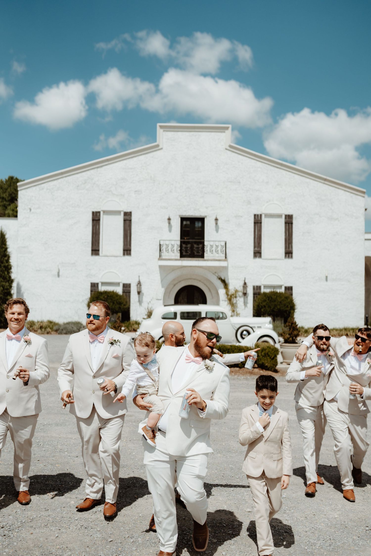 Groom and groomsmen in white suits celebrating outdoors in front of a white venue on a sunny wedding day.
