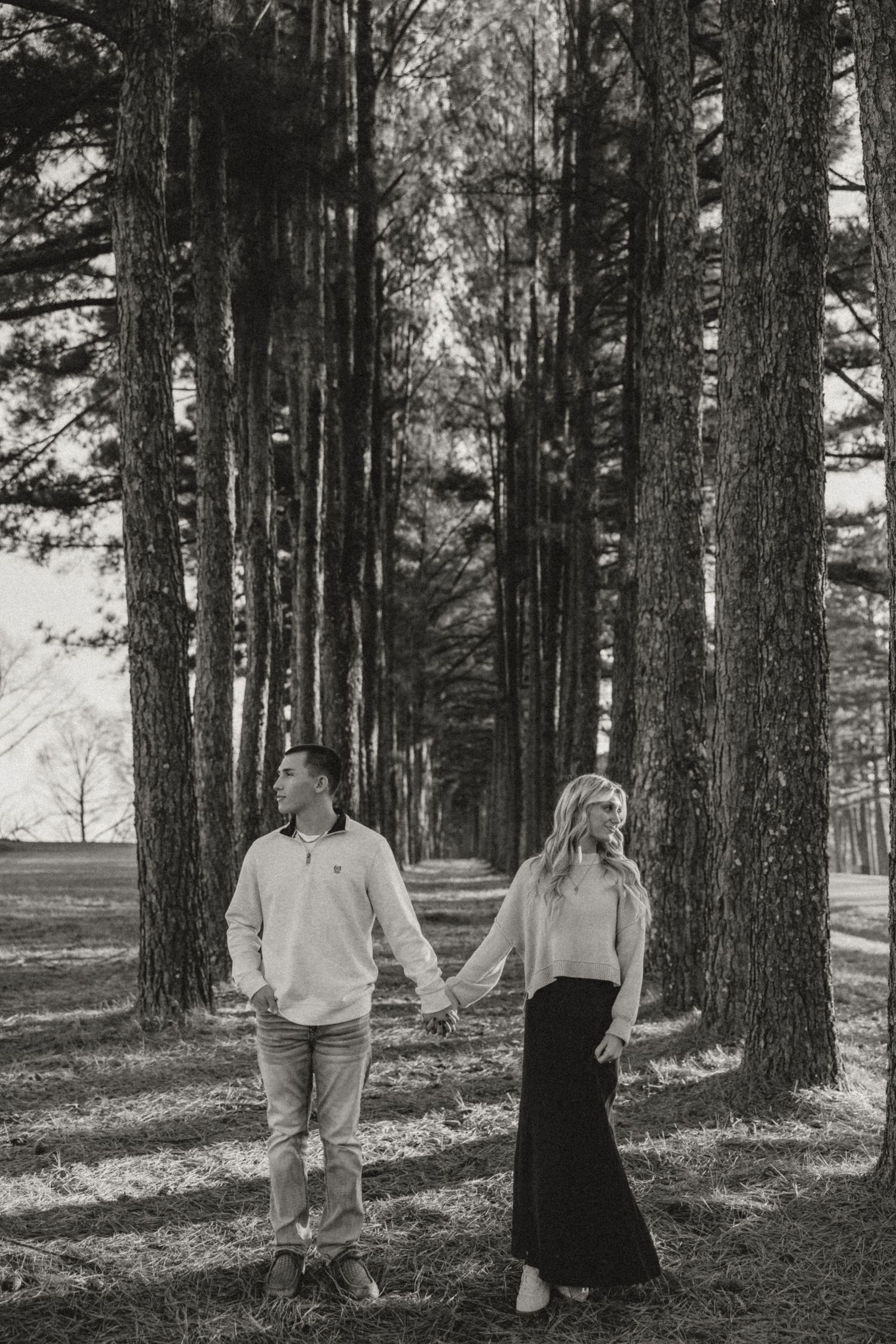 Black and white photo of a couple holding hands among tall pine trees in a serene forest setting.