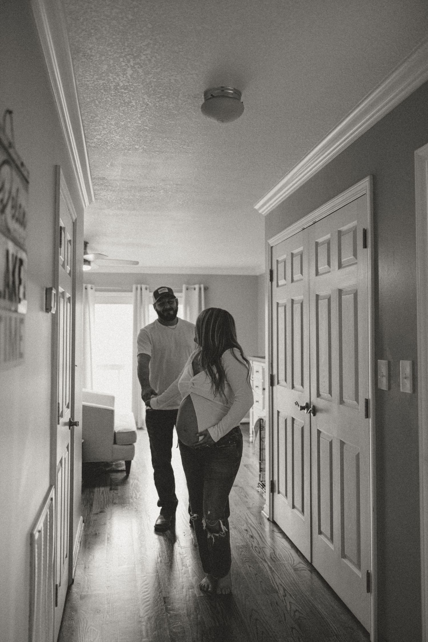 Black and white photo of a couple standing in a hallway, woman appears pregnant, sharing a tender moment at home.