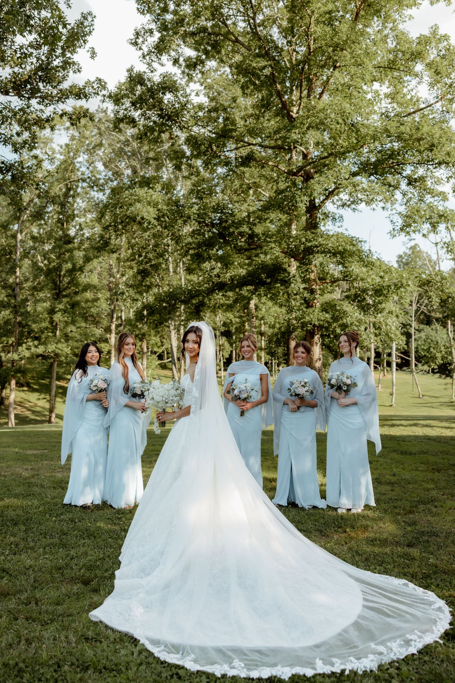 Bride in flowing gown with long train poses with six bridesmaids in light blue dresses in a park.