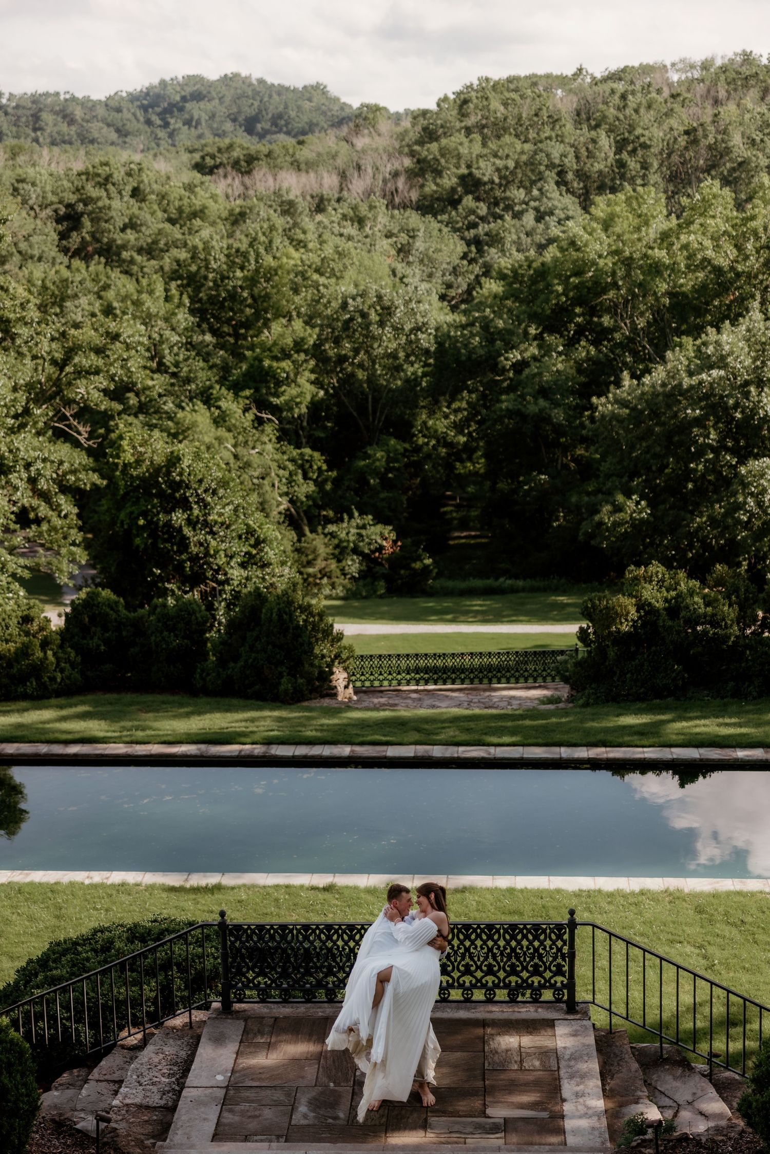 Two brides embrace on stone steps overlooking a serene reflecting pool surrounded by lush green trees.