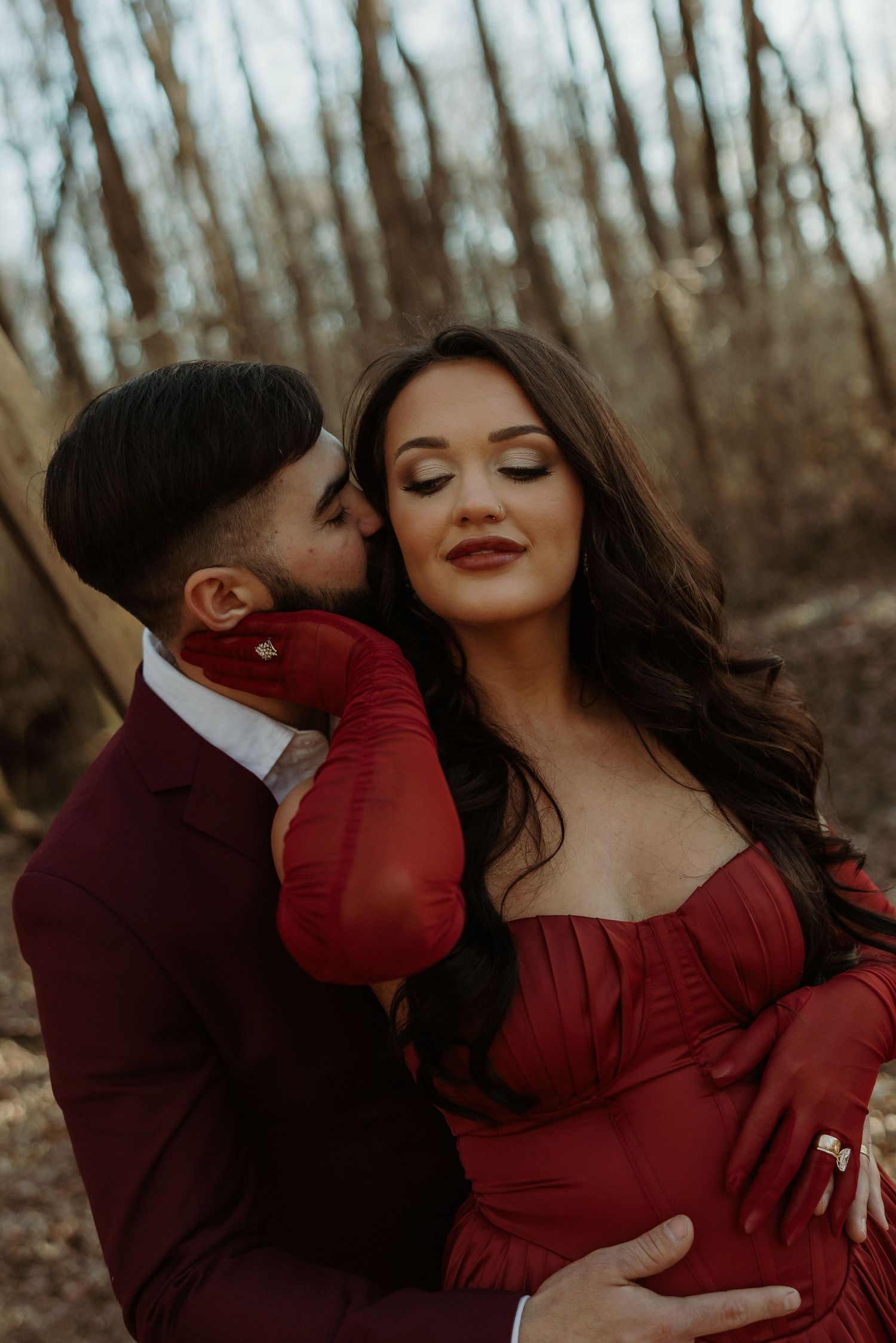 Couple in matching red outfits sharing a romantic moment outdoors, woman in off-shoulder gown cradling baby bump.