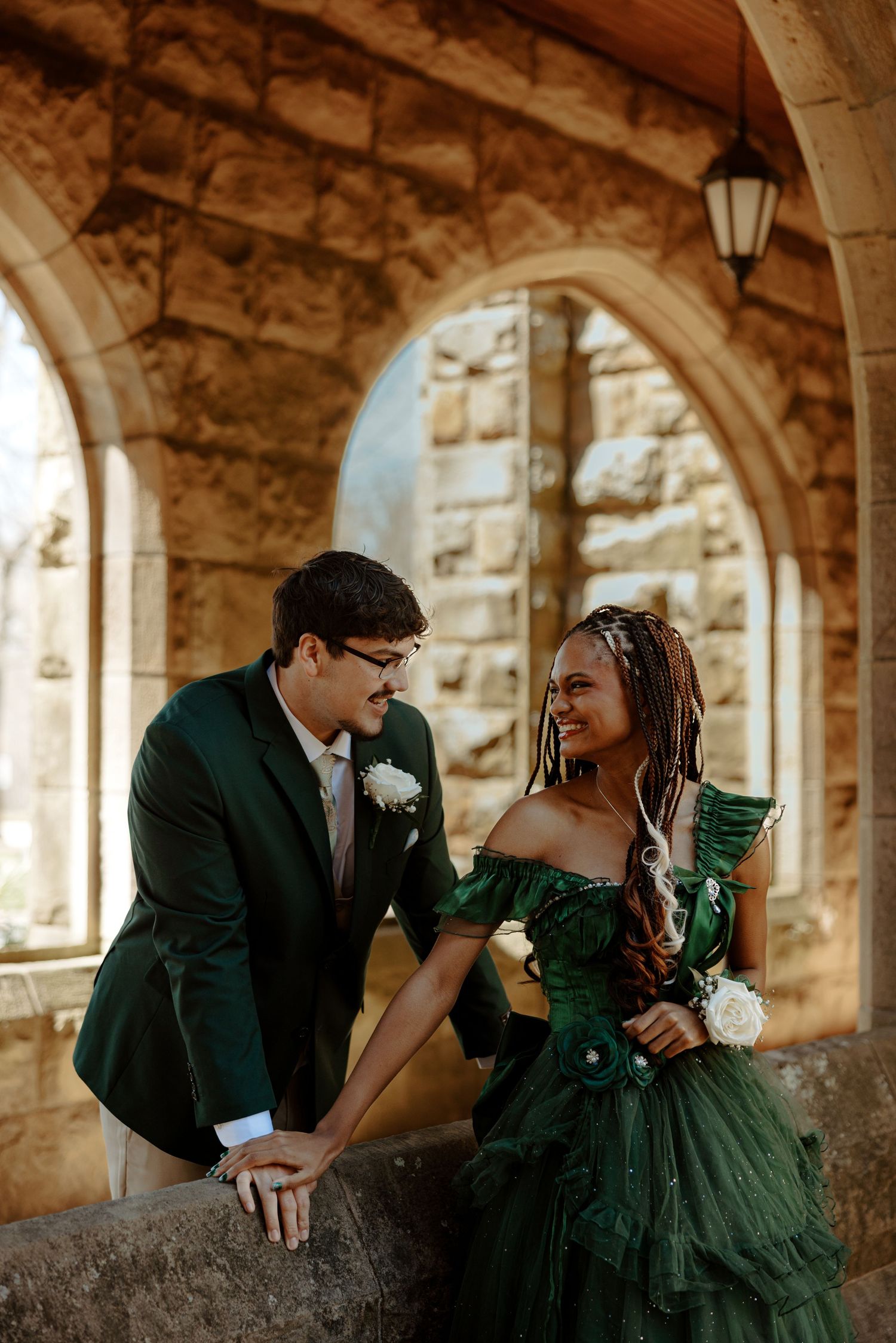 Couple in green formal wear posing under stone arches at elegant venue.