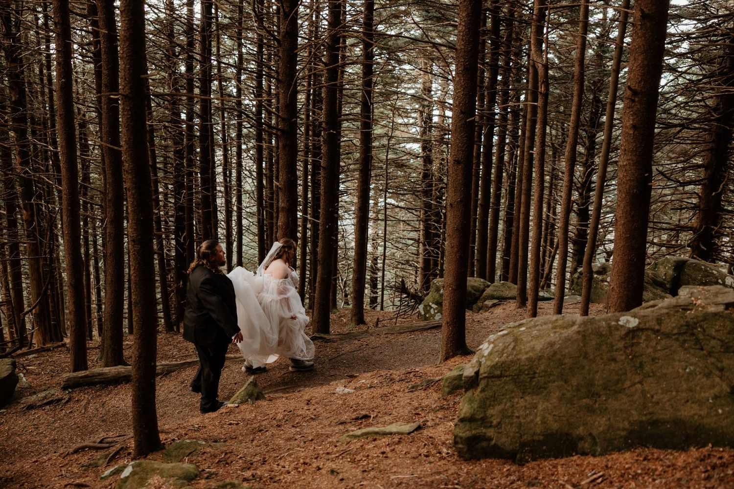 Bride and groom walking through a tall pine forest on their wedding day, with rocky terrain and autumn leaves.