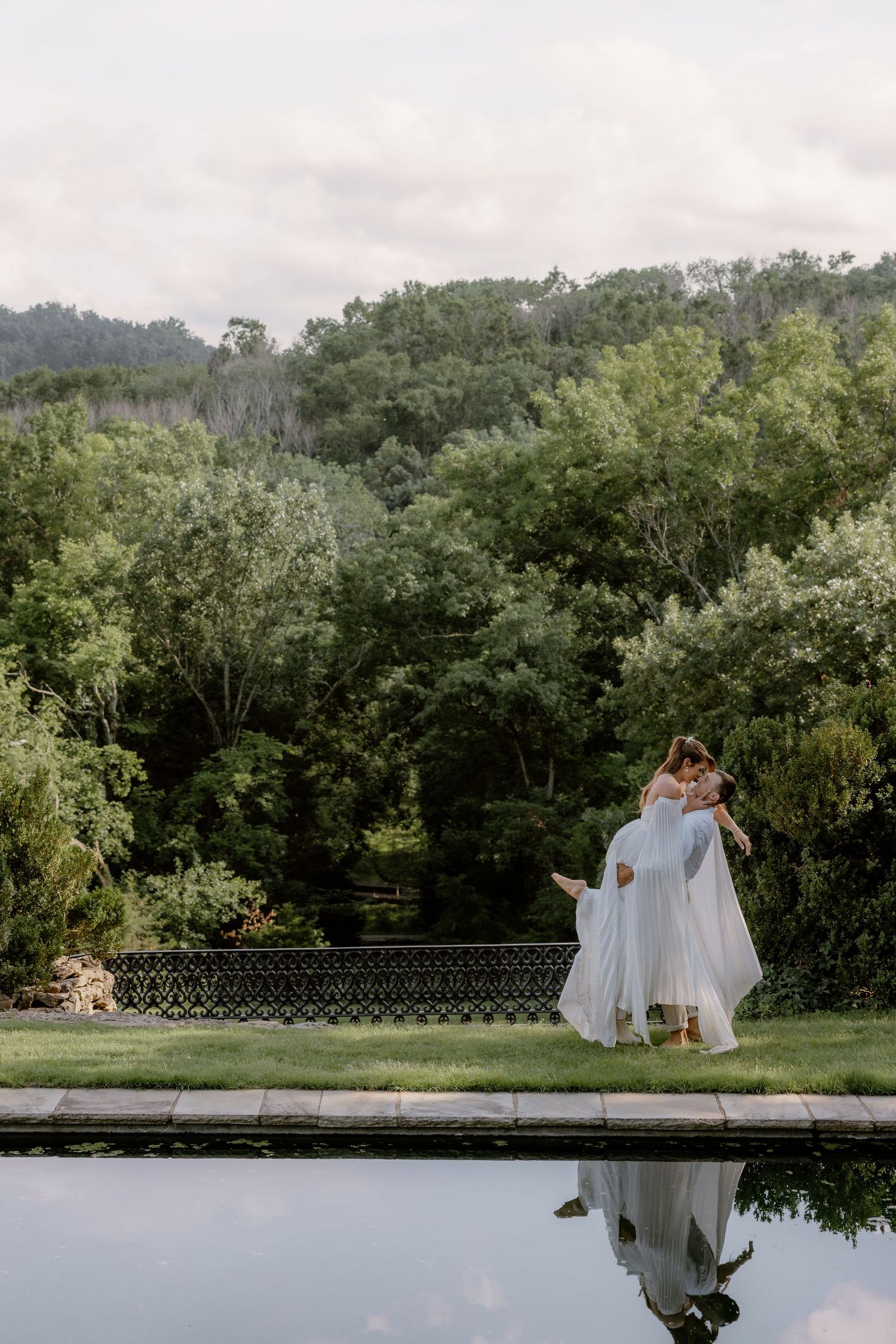 Couple embracing in wedding attire by a reflective pool with lush green forest backdrop on a cloudy day.