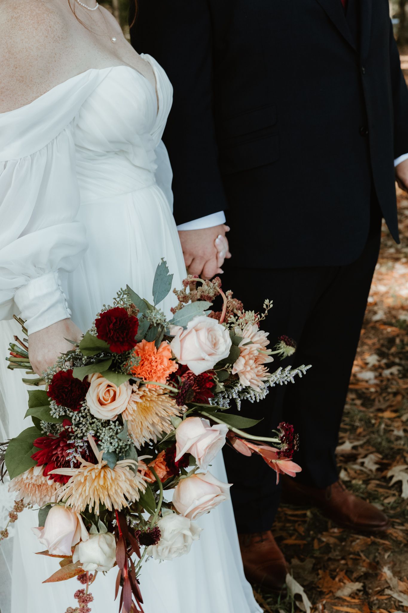 A close-up detail of a rustic fall wedding bouquet with burgundy and blush flowers against white dress and black suit.