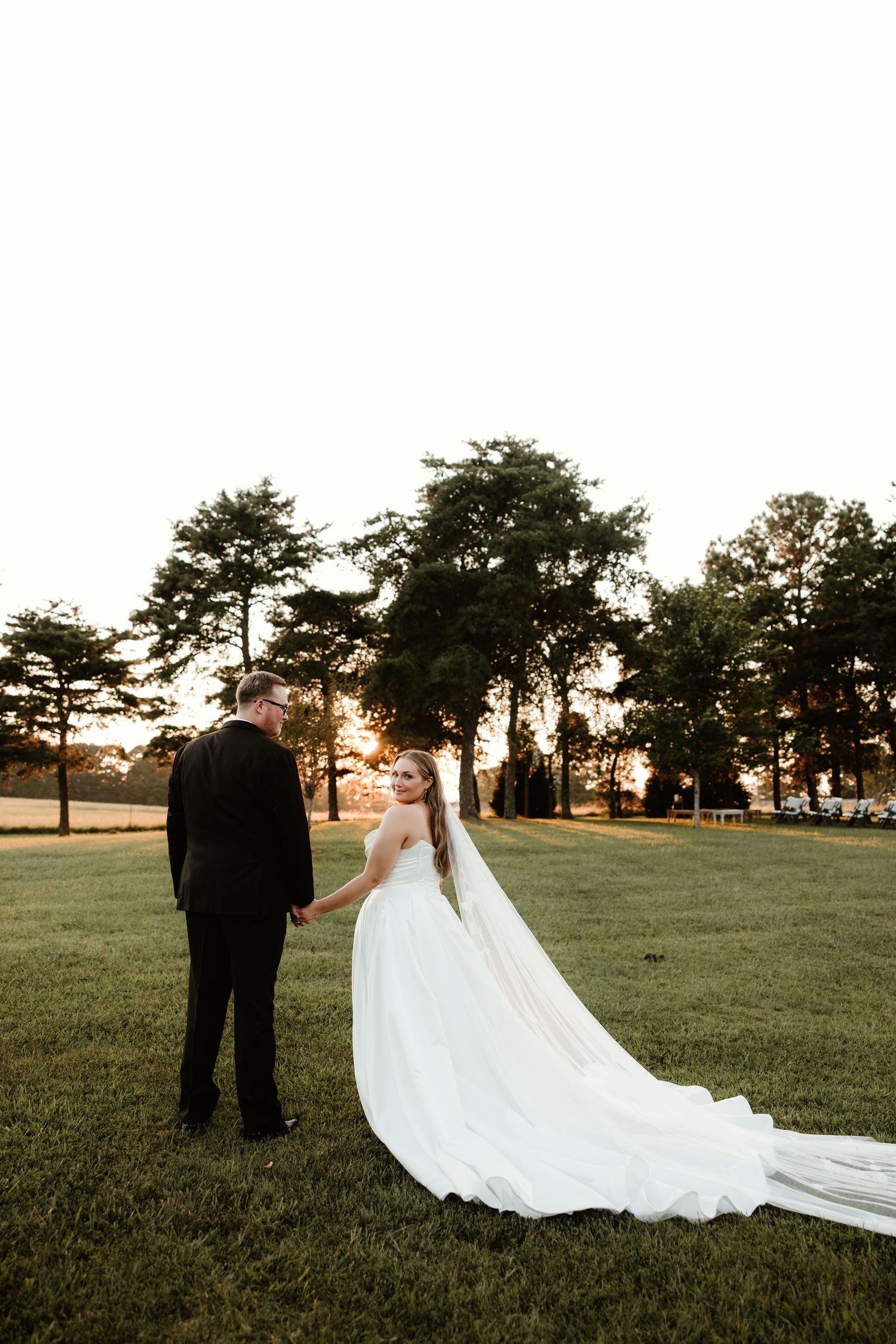 A couple in wedding attire stands in silhouette against the sunset on a grassy field during their outdoor ceremony.