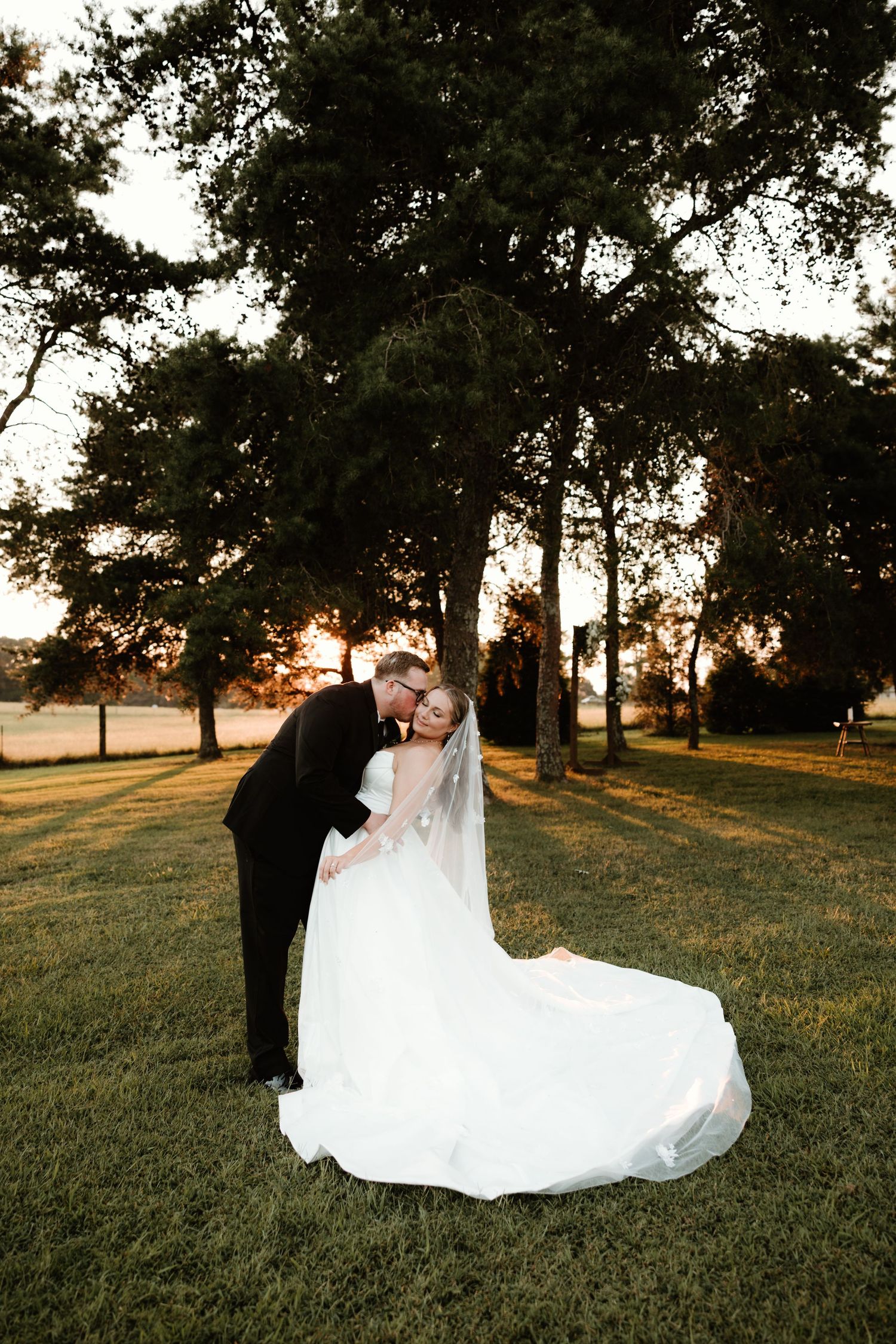 A romantic sunset wedding photo shows a couple sharing a kiss on a grassy lawn with tall trees silhouetted against the sky.
