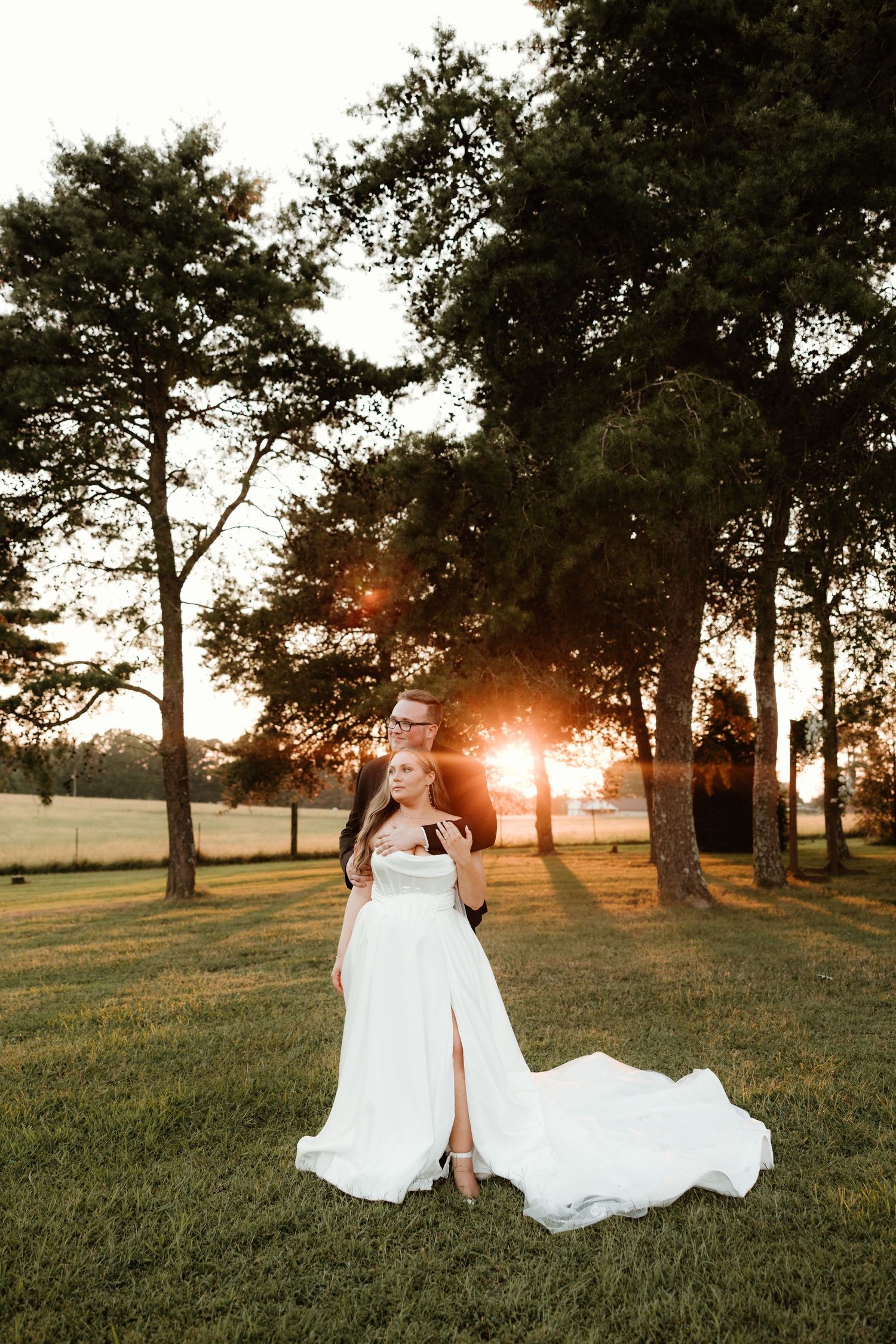 A stunning sunset wedding portrait featuring a white flowing dress with a slit on a grassy lawn framed by tall trees.