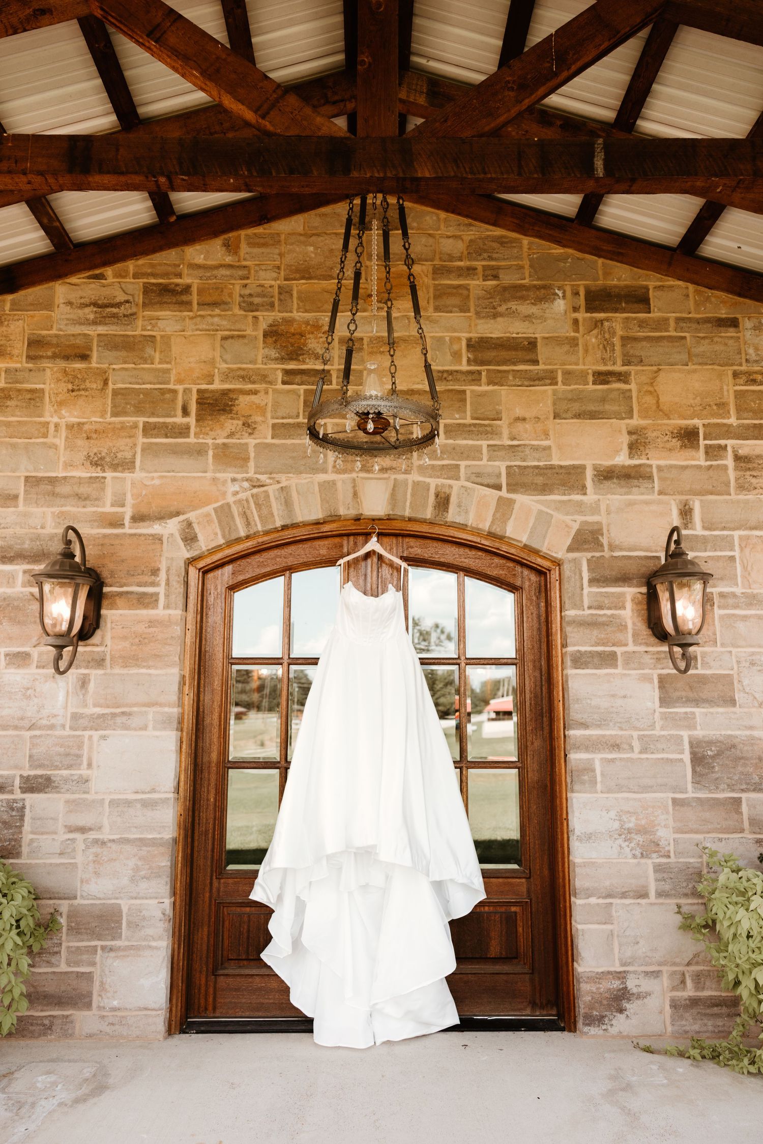 A white wedding dress hangs on display in front of an elegant stone archway with wooden doors and wall sconces.