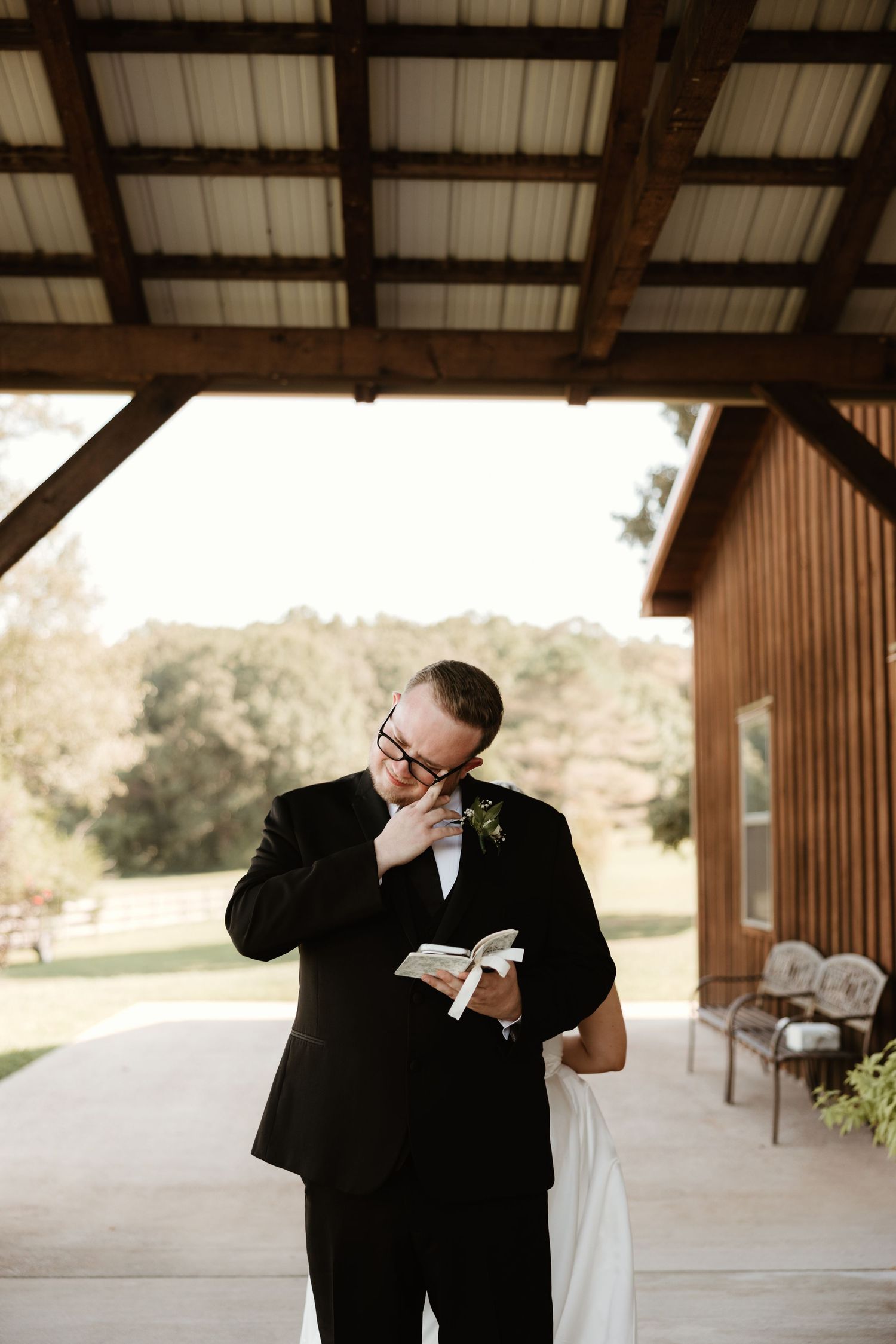 An emotional moment captured at a rustic outdoor venue as someone in a black suit reads from a small book.