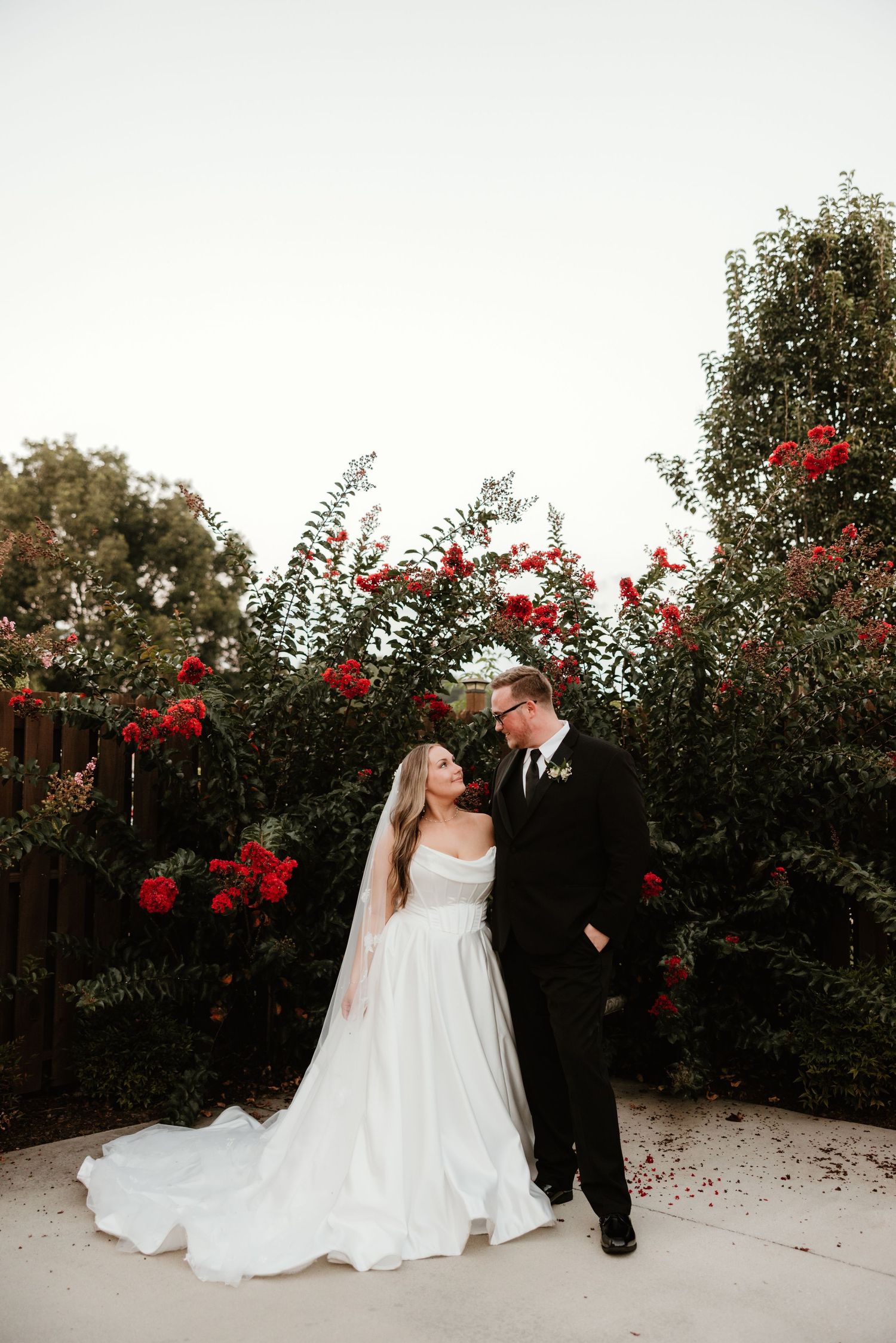 A romantic newlywed couple stands together in a garden with blooming red roses and lush greenery during sunset.