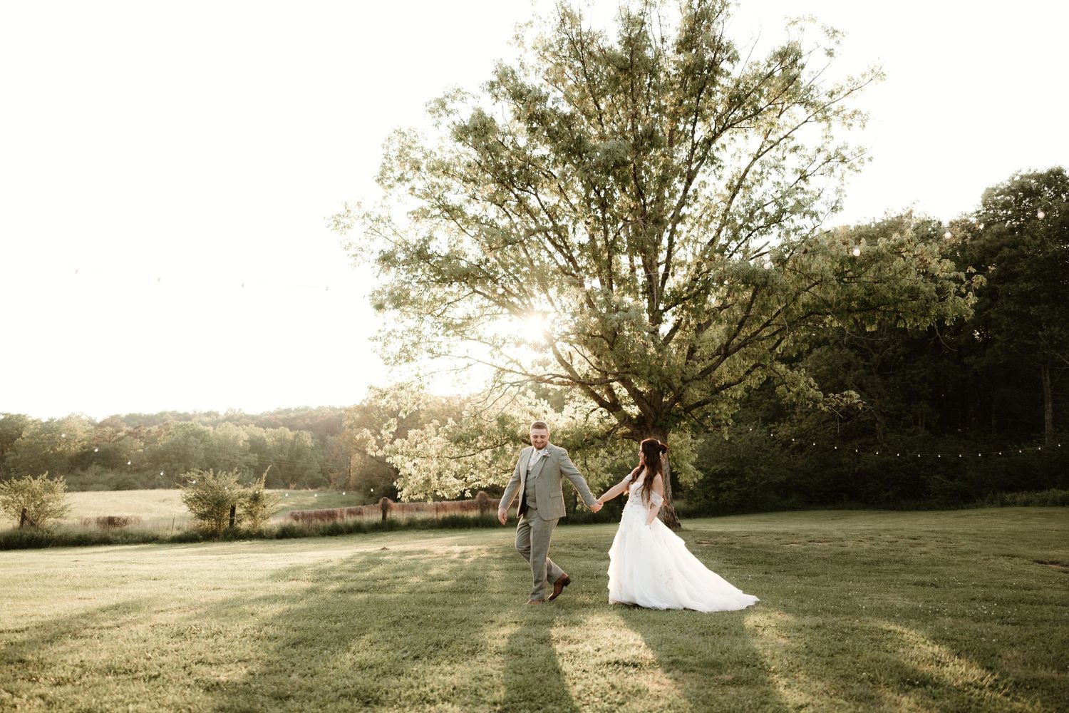 A couple in wedding attire stands beneath a sunlit tree on a rolling grassy hillside at sunset.