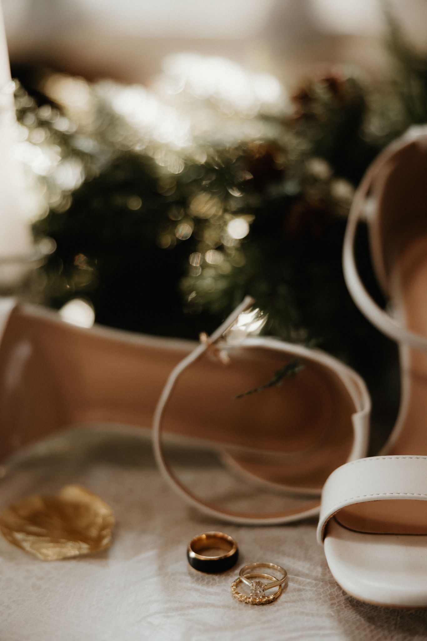 Wedding rings and white bridal shoes arranged with greenery in soft, romantic lighting.