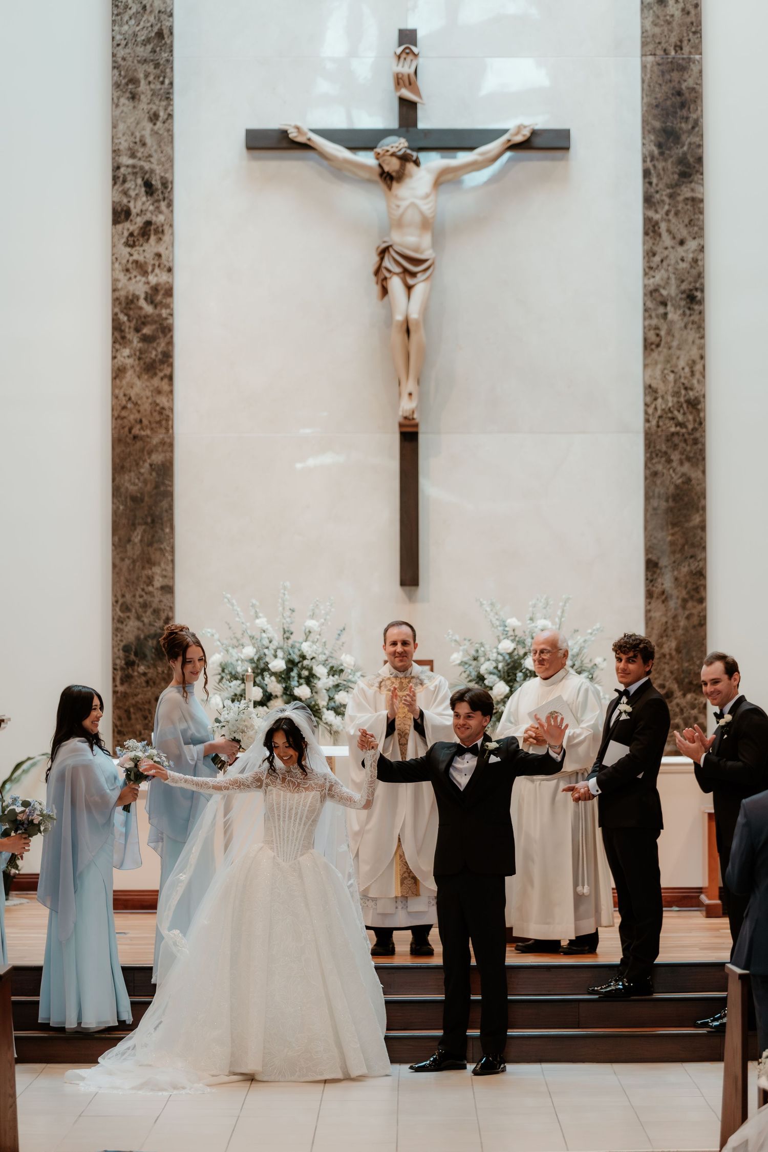 Newlyweds raise joined hands in celebration at church altar beneath crucifix with wedding party applauding.