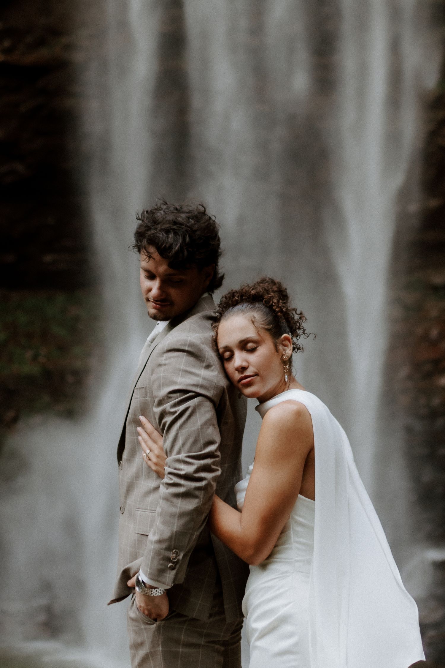 A bride in a white gown leans on a groom in a gray suit, standing before a misty waterfall backdrop.
