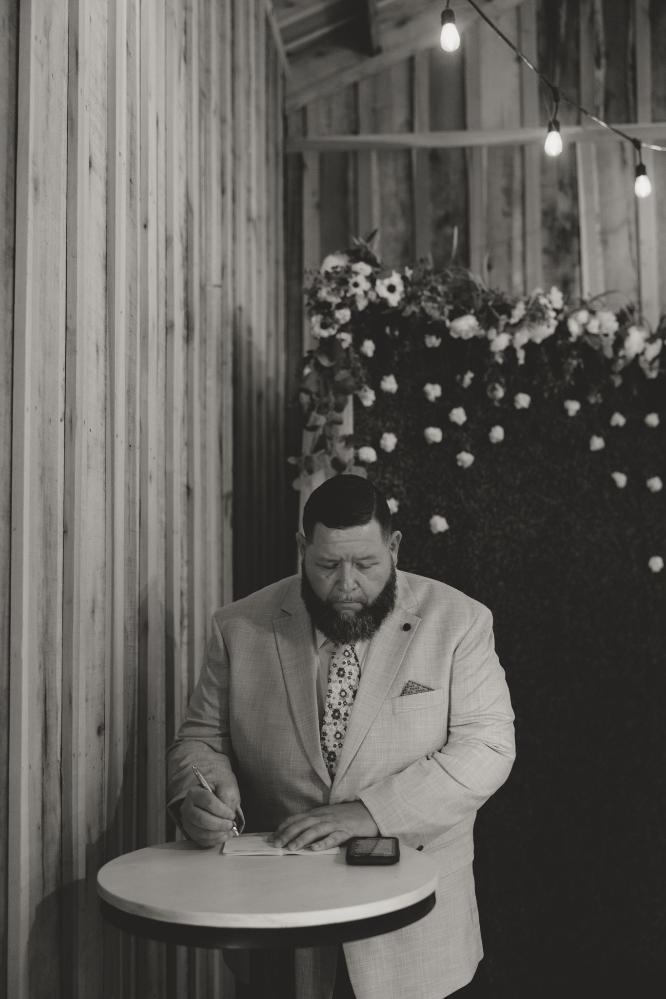Person in light suit signing documents at round table with flower decorations and string lights in rustic wooden venue.