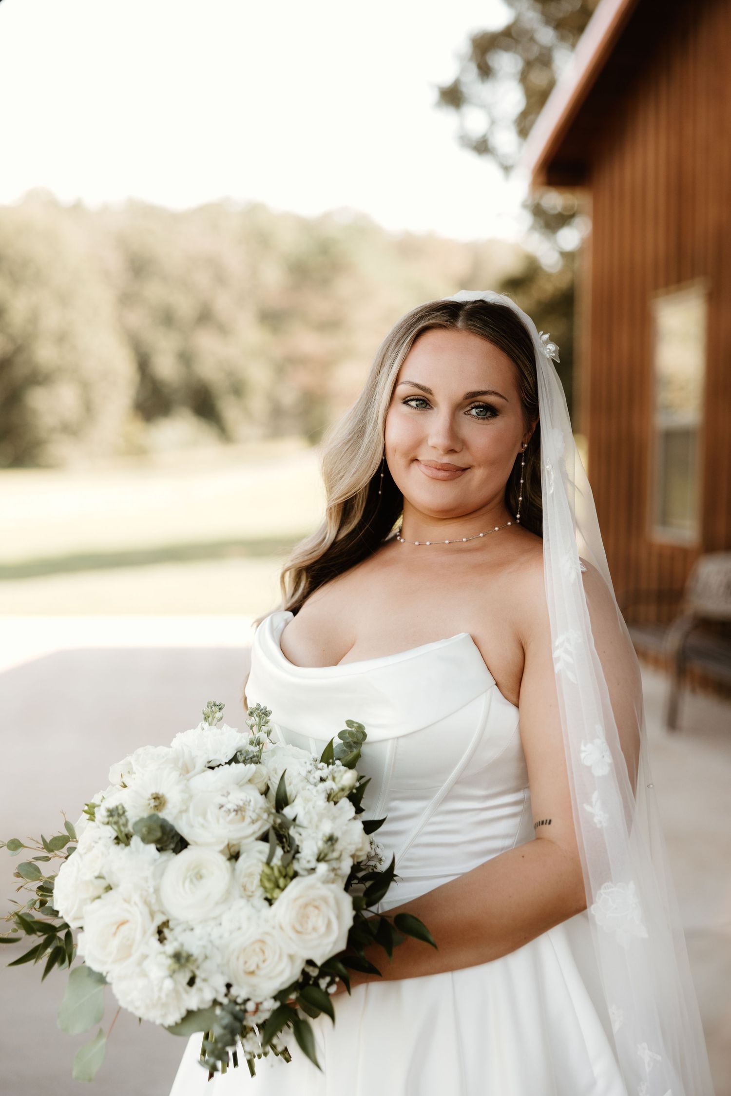 Beautiful bride in white wedding dress poses with elegant white and green floral bouquet outside rustic venue.