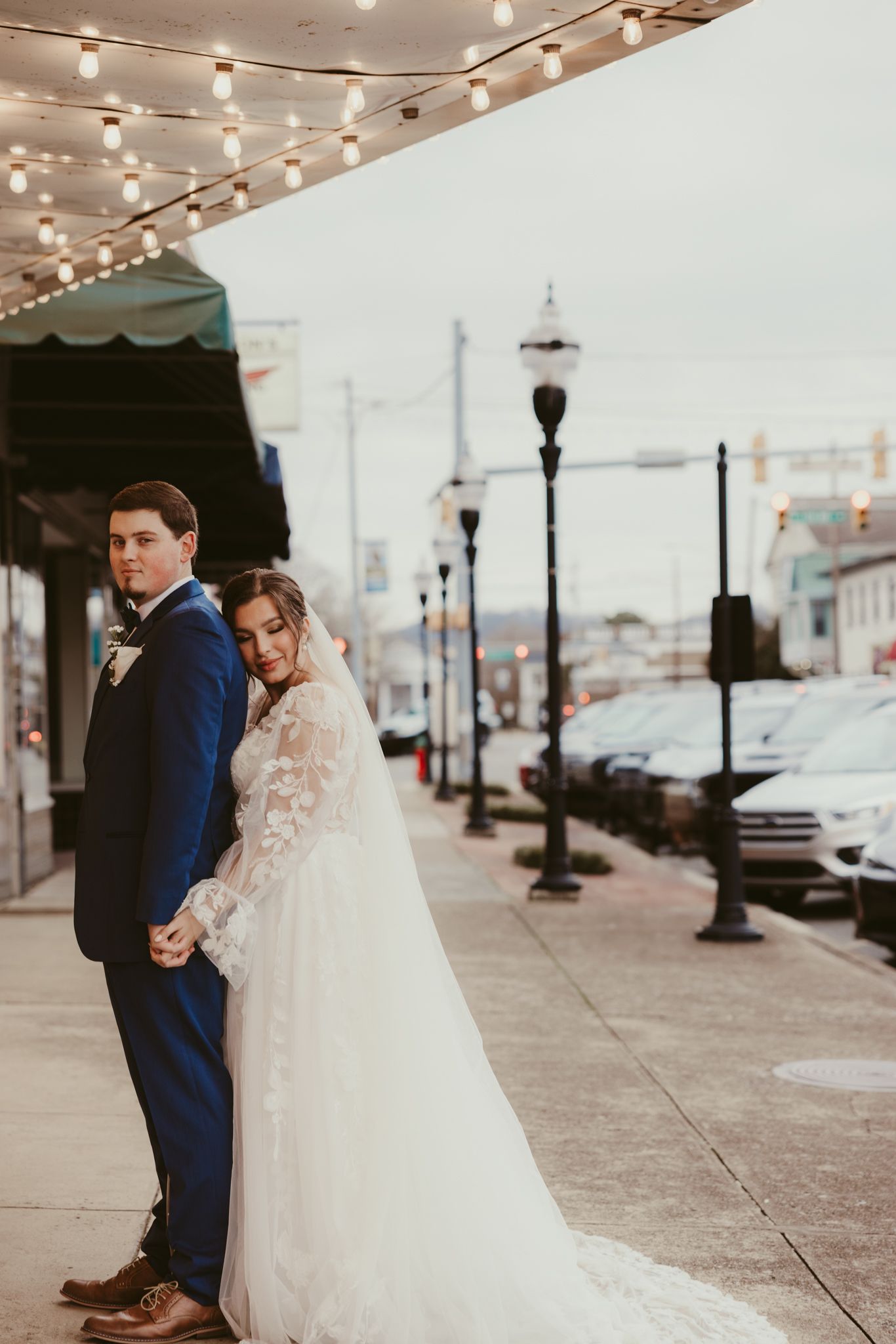 A couple poses under string lights on a city sidewalk with a long flowing wedding veil.