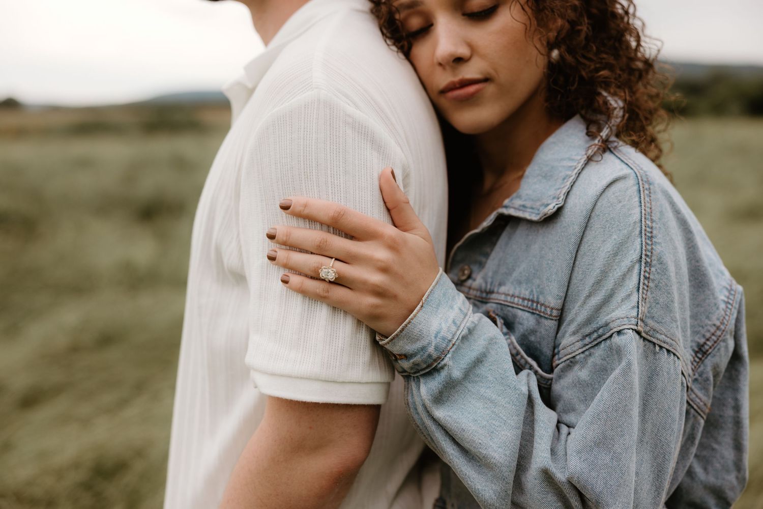 A person in a denim jacket rests their head gently on their partner's shoulder in a green field.