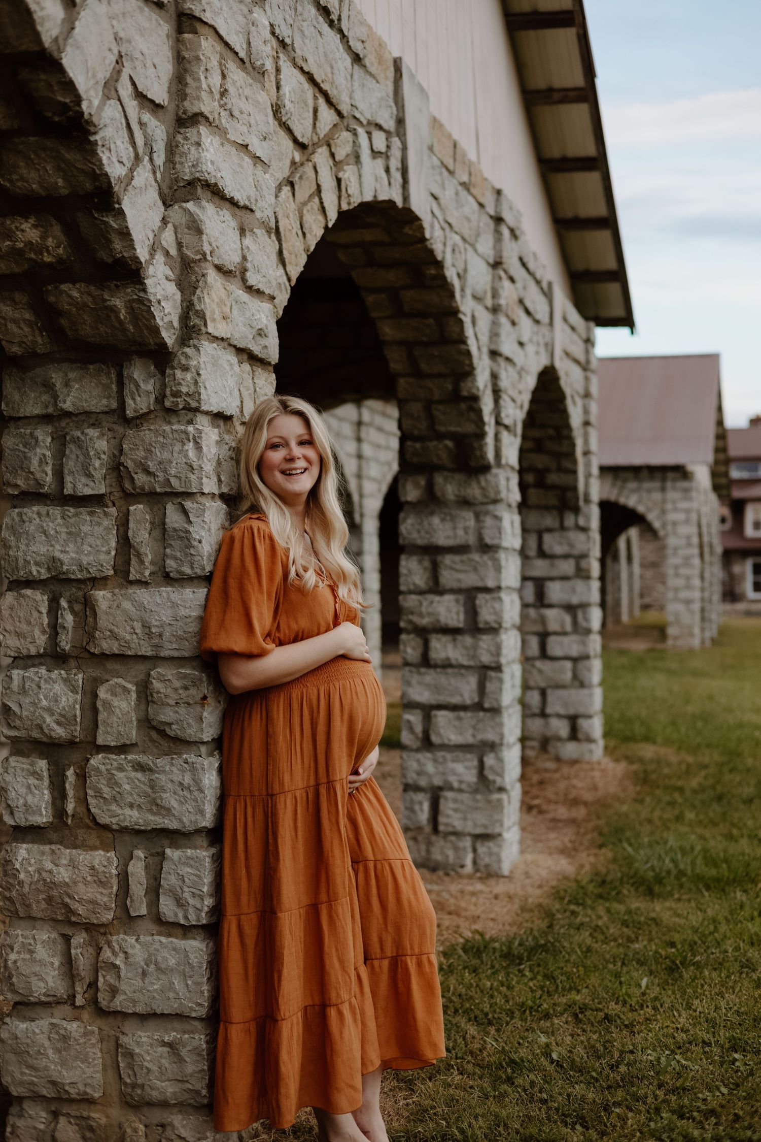 A woman in a flowing burnt orange tiered maxi dress poses against a stone archway outdoors during golden hour.