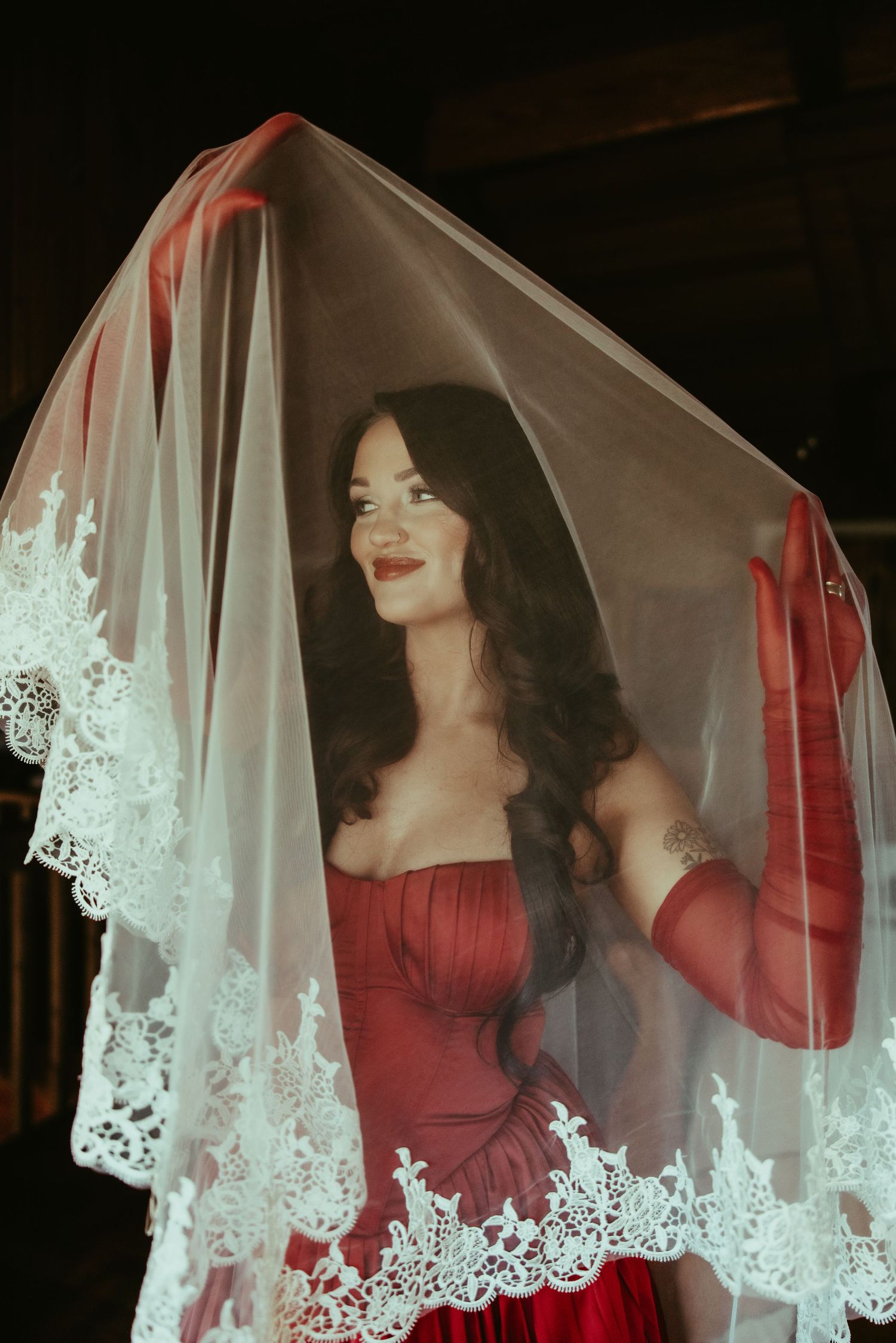 Woman in red strapless gown smiling under white lace bridal veil in dramatic dark setting.